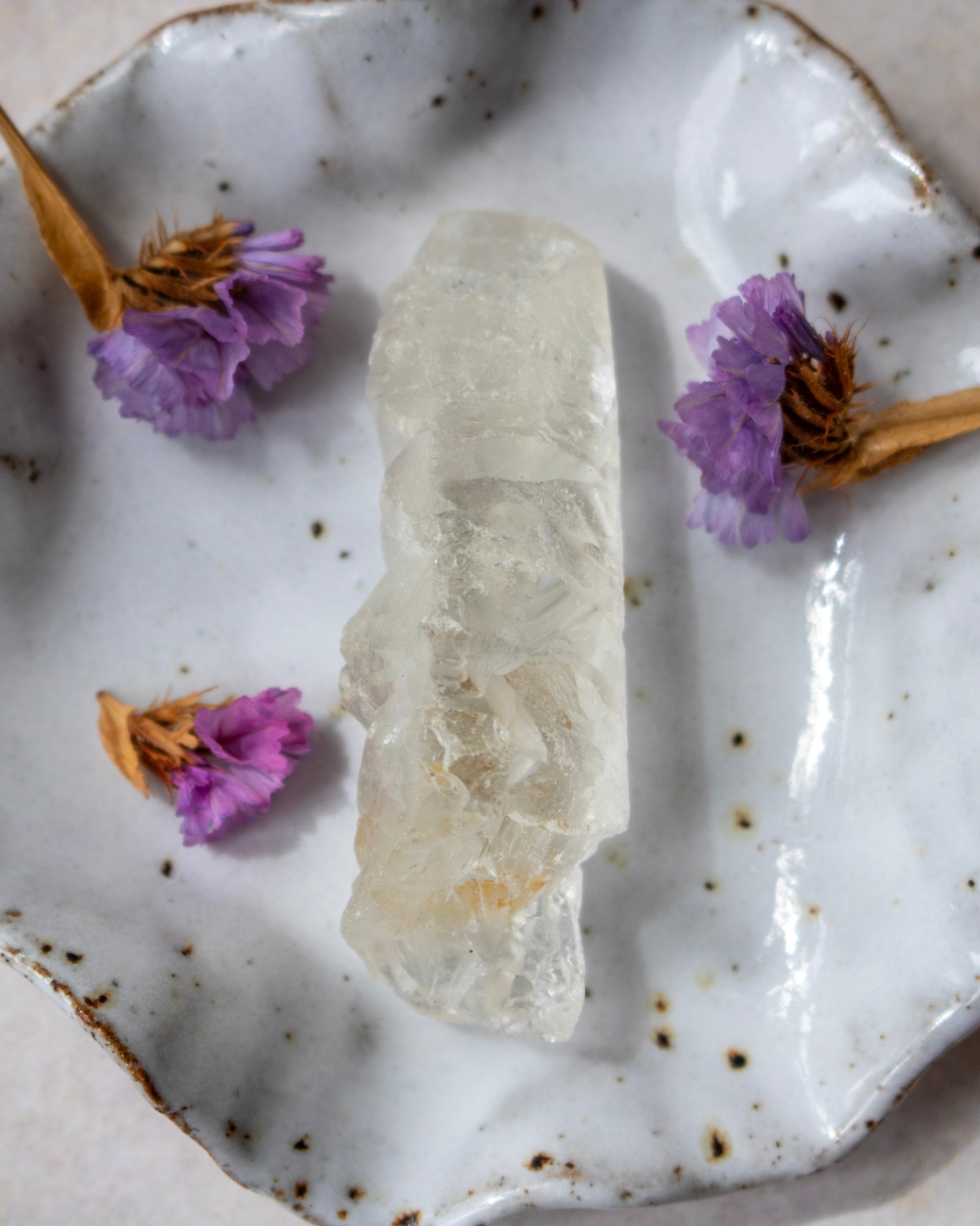 A natural clear quartz crystal cluster sits in the center of a white speckled ceramic dish decorated with dried purple flowers.