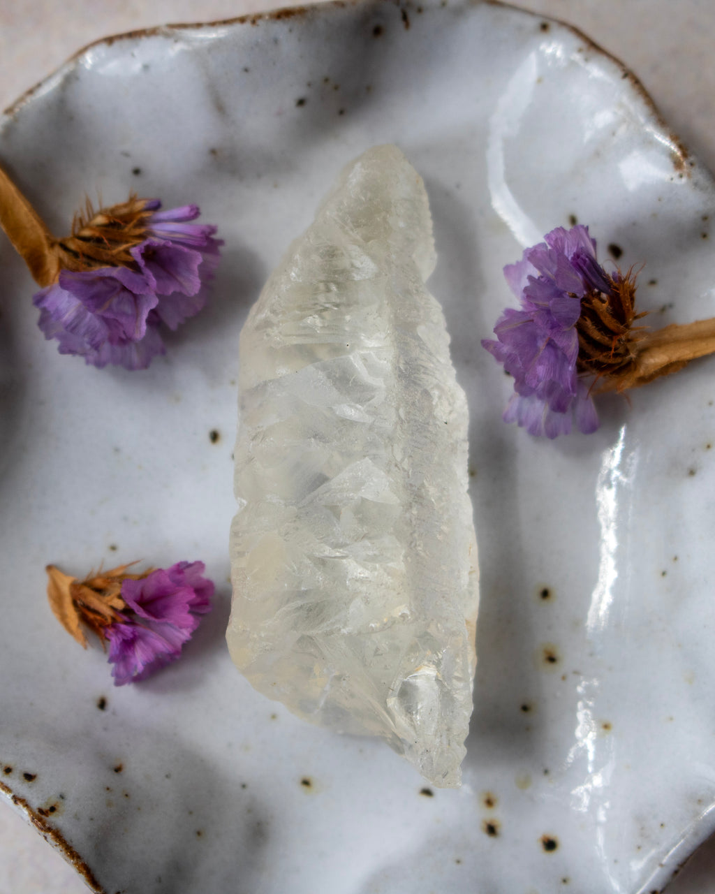A raw, translucent pale crystal stone placed on a speckled white ceramic plate with dried purple flowers around it.