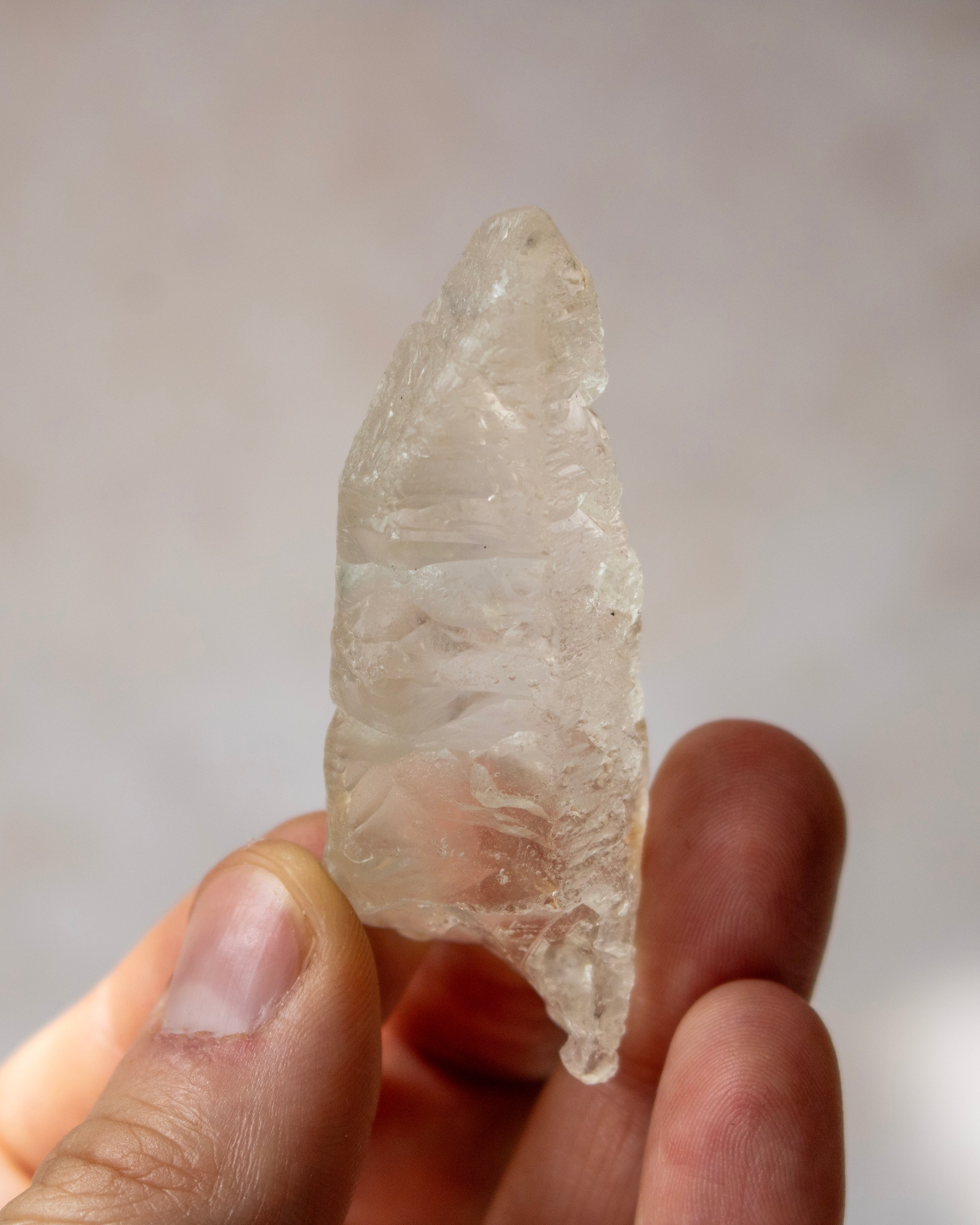 A hand holding a raw, naturally faceted clear quartz crystal point against a soft, out-of-focus light gray background.