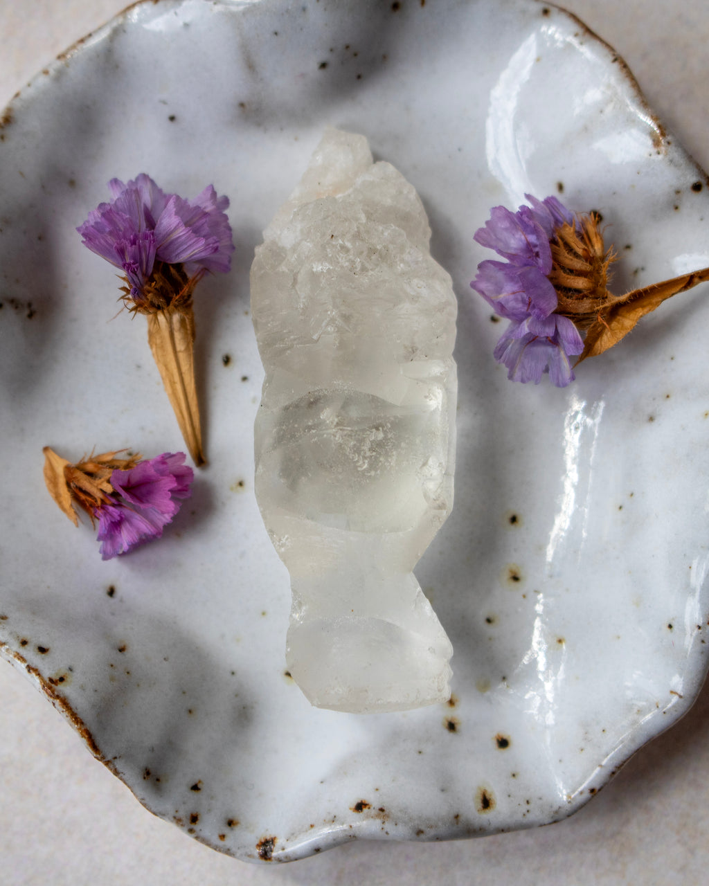 A raw, rough-cut clear quartz crystal cluster displayed on a rustic, speckled white ceramic plate with dried purple flowers.