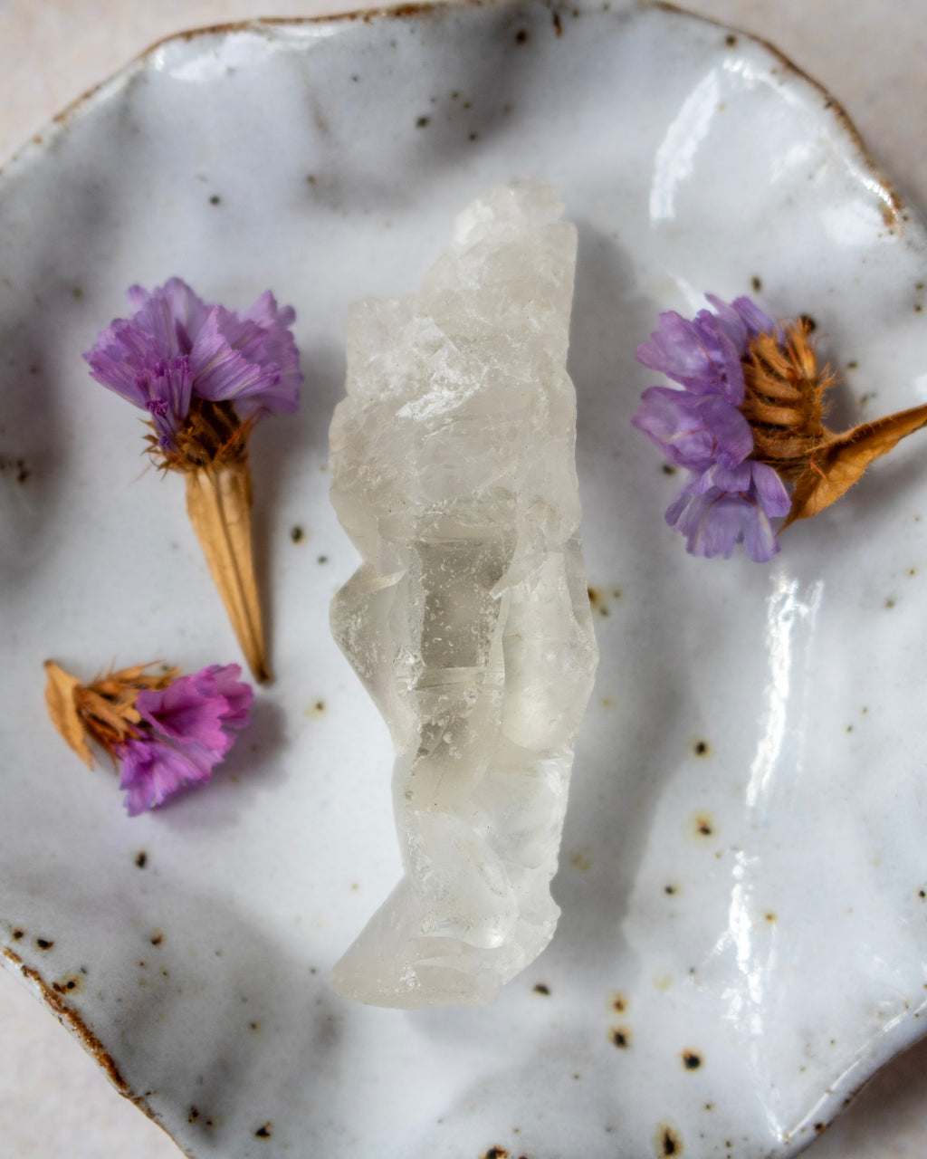 A polished white crystal cluster, likely quartz, displayed on a rustic white speckled ceramic plate with dried purple flowers as decorative props.