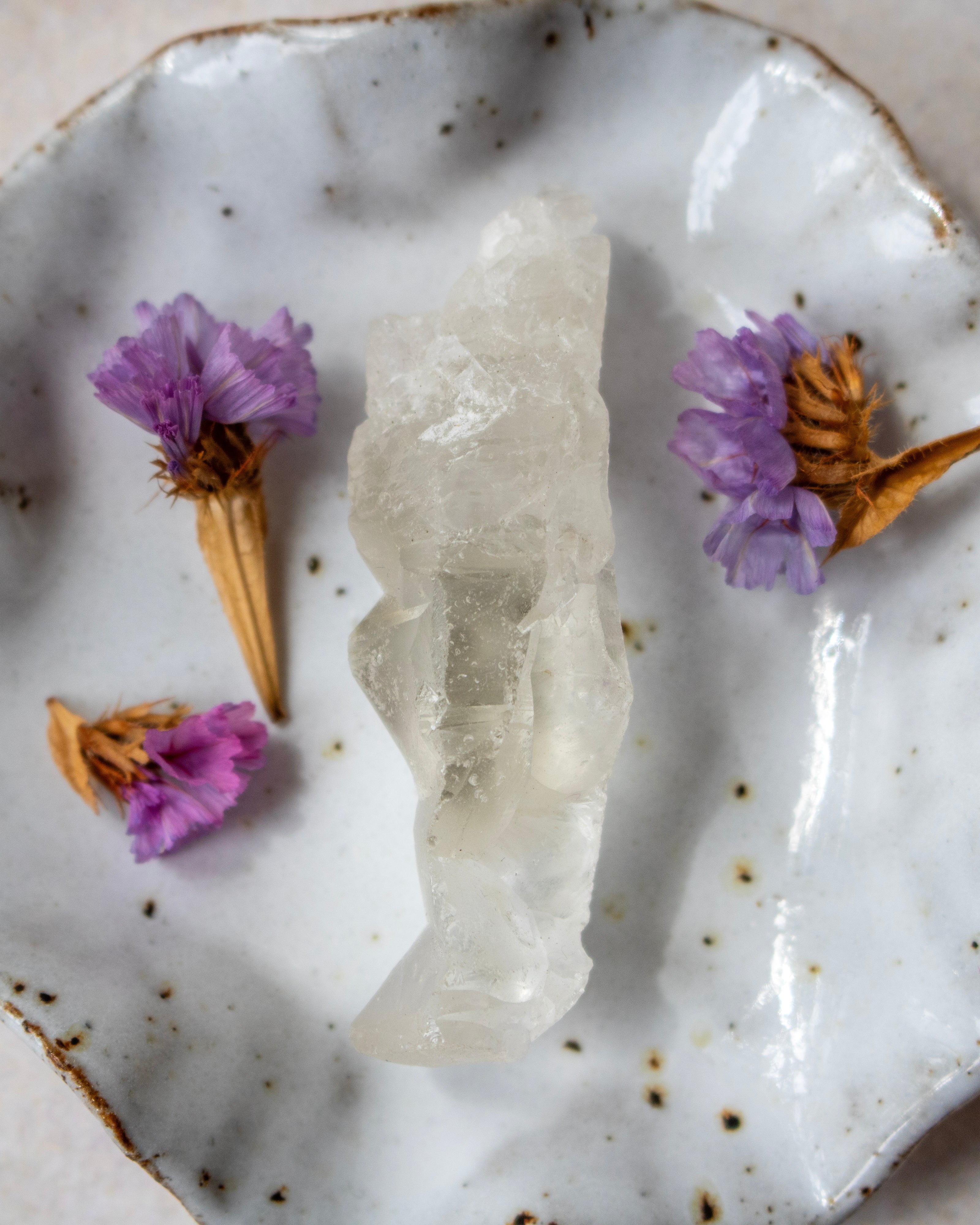 A polished white crystal cluster, likely quartz, displayed on a rustic white speckled ceramic plate with dried purple flowers as decorative props.