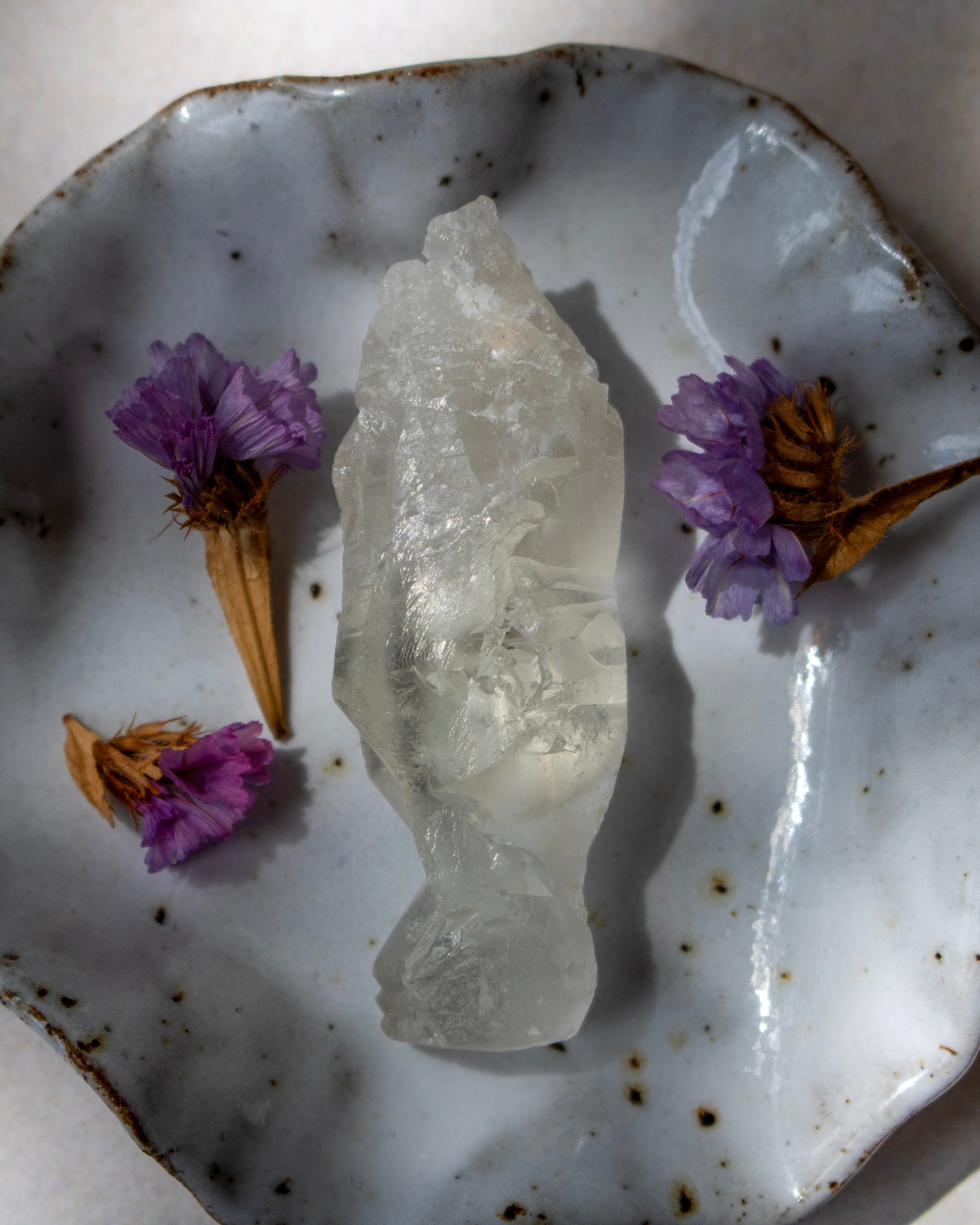 A rough, unpolished clear quartz crystal cluster sits in the center of a speckled white ceramic dish, styled with small dried purple flowers on a light beige surface.