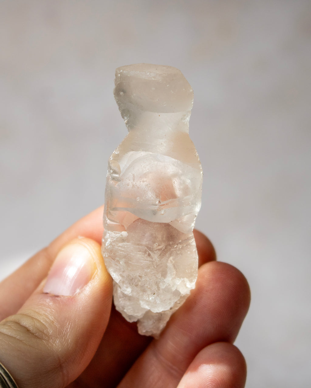 A hand holding a small, rough, translucent pale crystal or stone figurine against a soft light gray background.