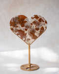 Heart-shaped crystal with a brown and white agate-like pattern, mounted on a gold metal stand against a neutral light gray background.