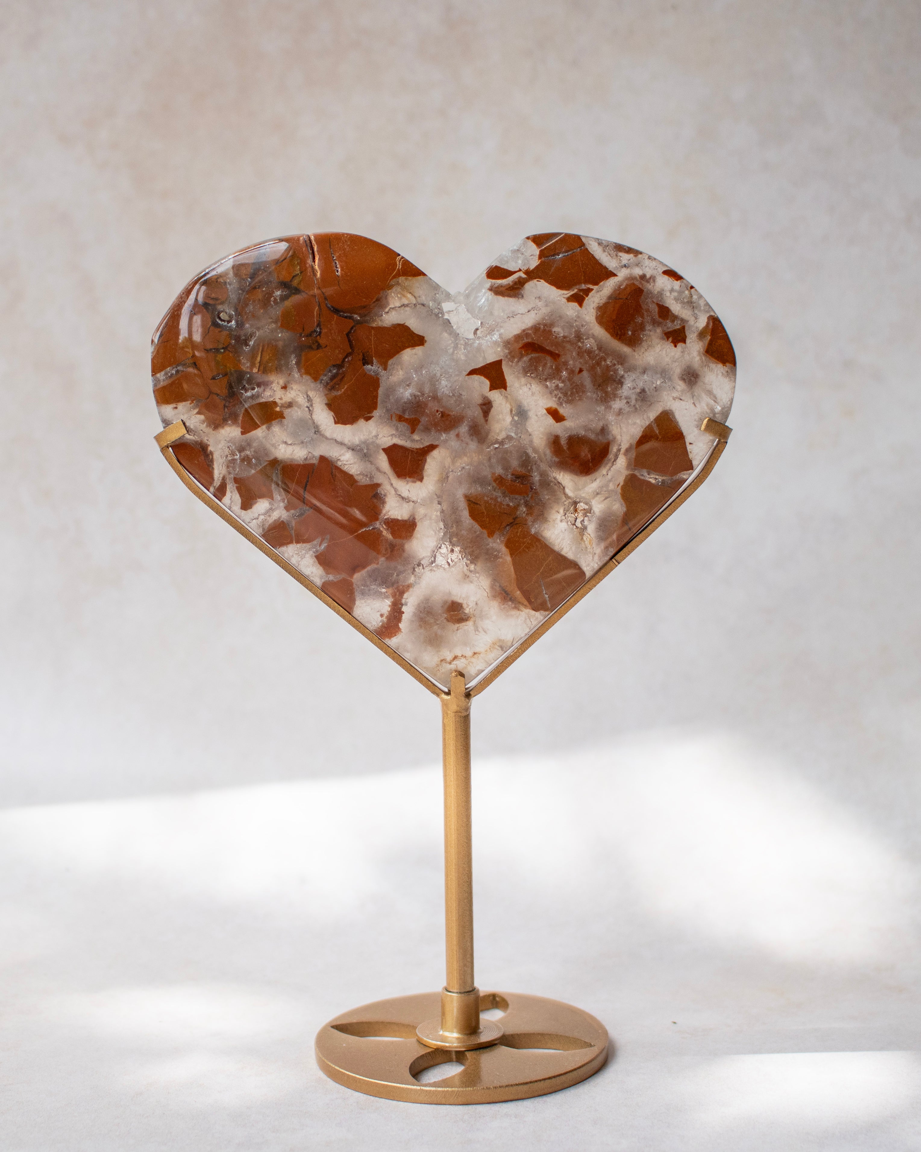Heart-shaped crystal with a brown and white agate-like pattern, mounted on a gold metal stand against a neutral light gray background.