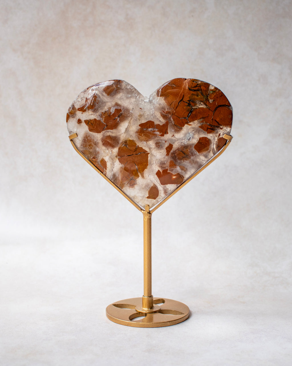 A heart-shaped crystal with a brown and white marbled pattern, mounted on a polished gold metal stand against a neutral textured background.