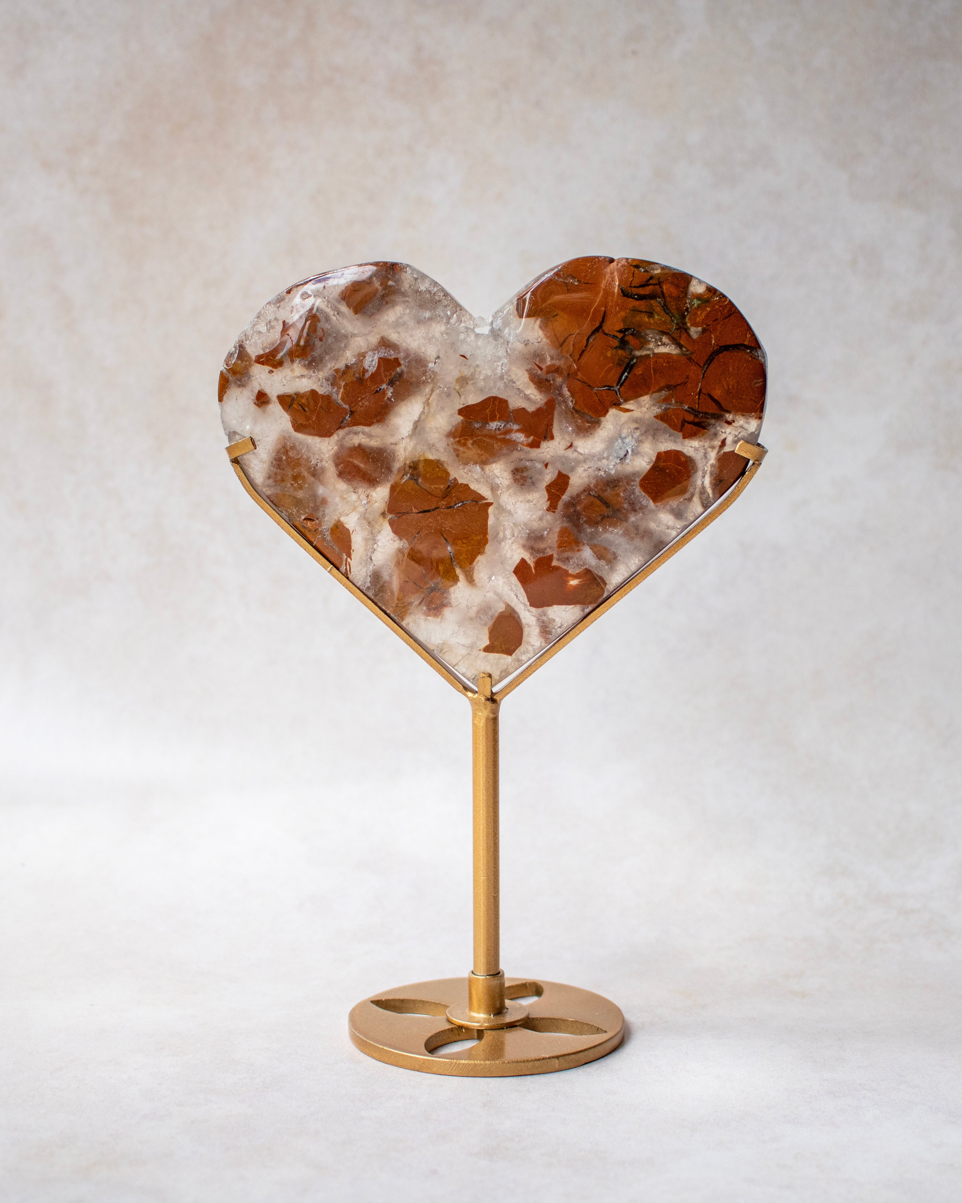 A heart-shaped crystal with a brown and white marbled pattern, mounted on a polished gold metal stand against a neutral textured background.