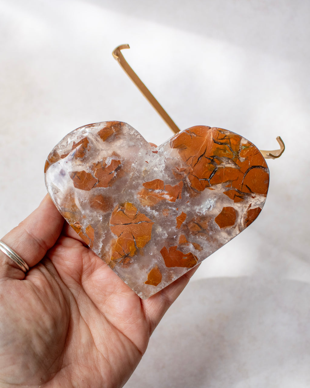 A heart-shaped crystal cluster with orange and brown mineral flecks, held in a hand against a soft white background.