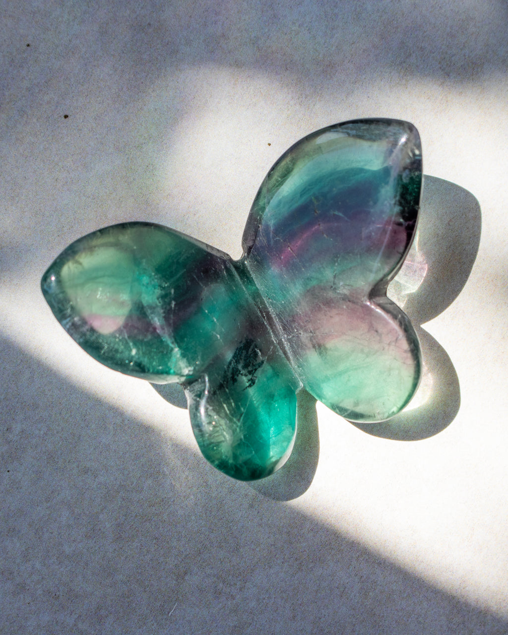 A butterfly-shaped translucent glass object with iridescent green, teal, and purple-blue tones, photographed on a light surface with strong natural shadows.