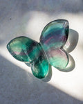 A butterfly-shaped translucent glass object with iridescent green, teal, and purple-blue tones, photographed on a light surface with strong natural shadows.