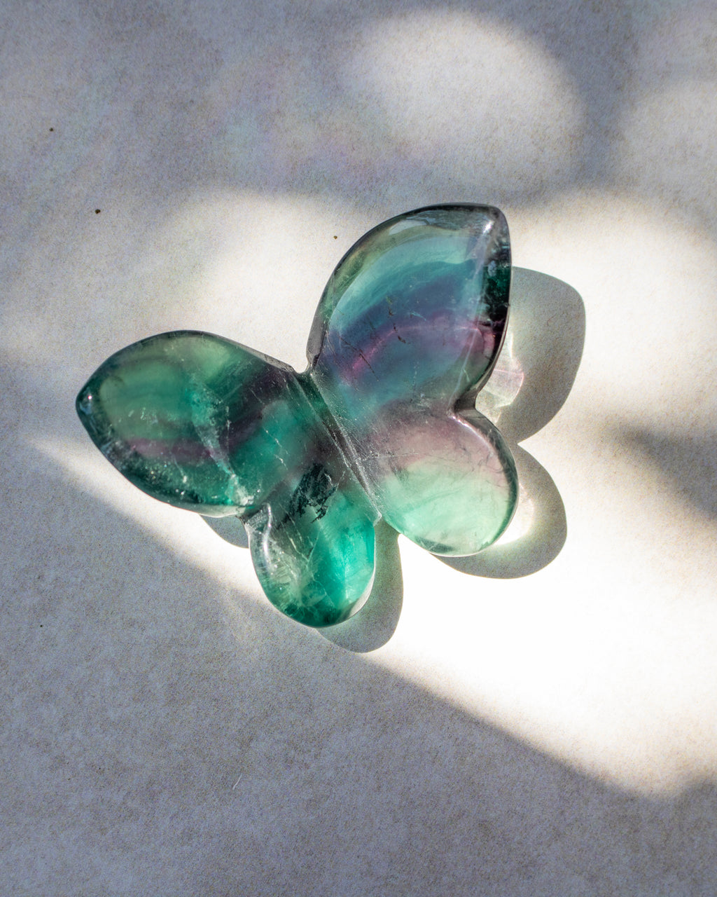 A butterfly-shaped glass art piece with iridescent green, blue, and purple hues, resting on a light textured surface with dappled sunlight and shadows.