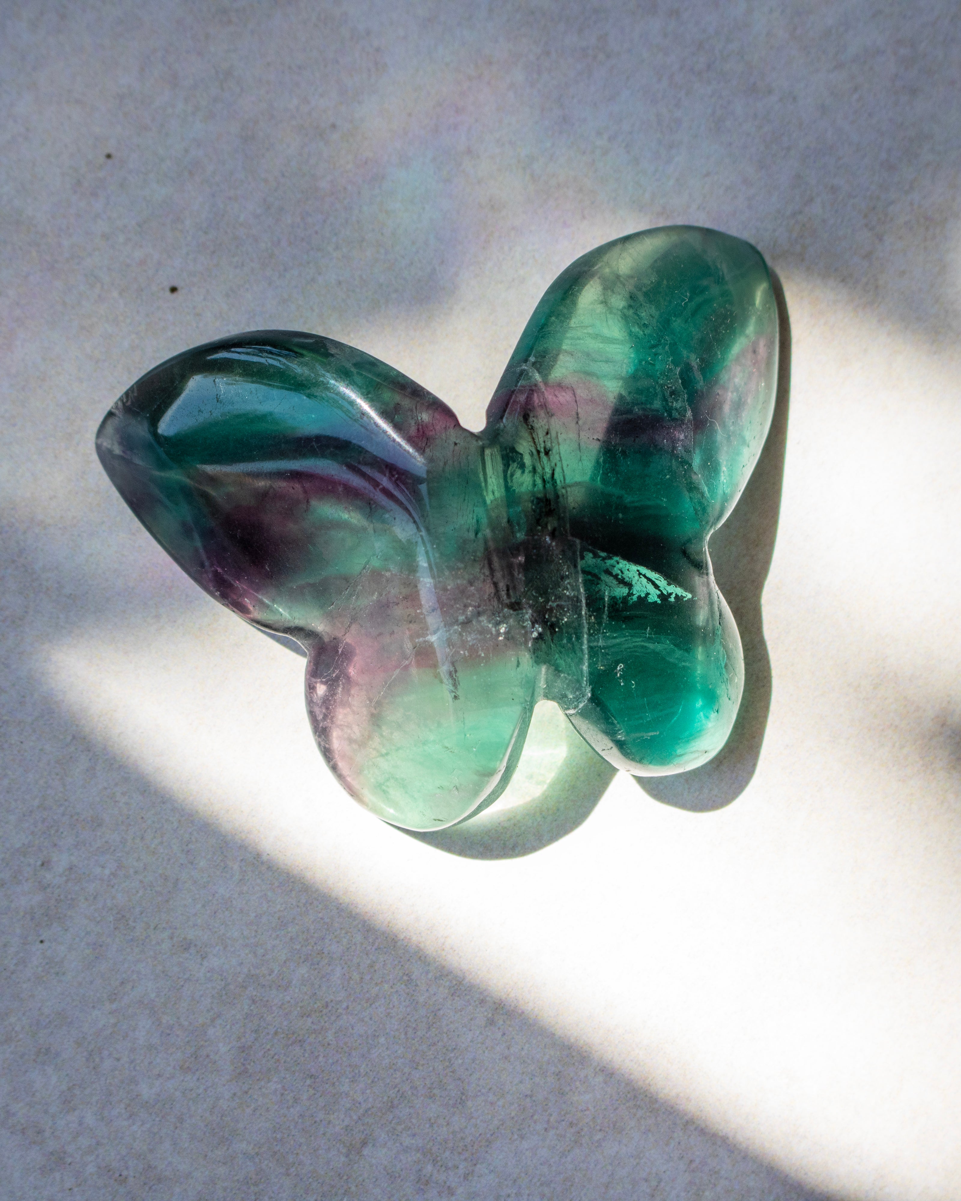 A translucent butterfly-shaped glass sculpture with green and purple marbling, photographed on a light surface with strong sunlight and shadows.
