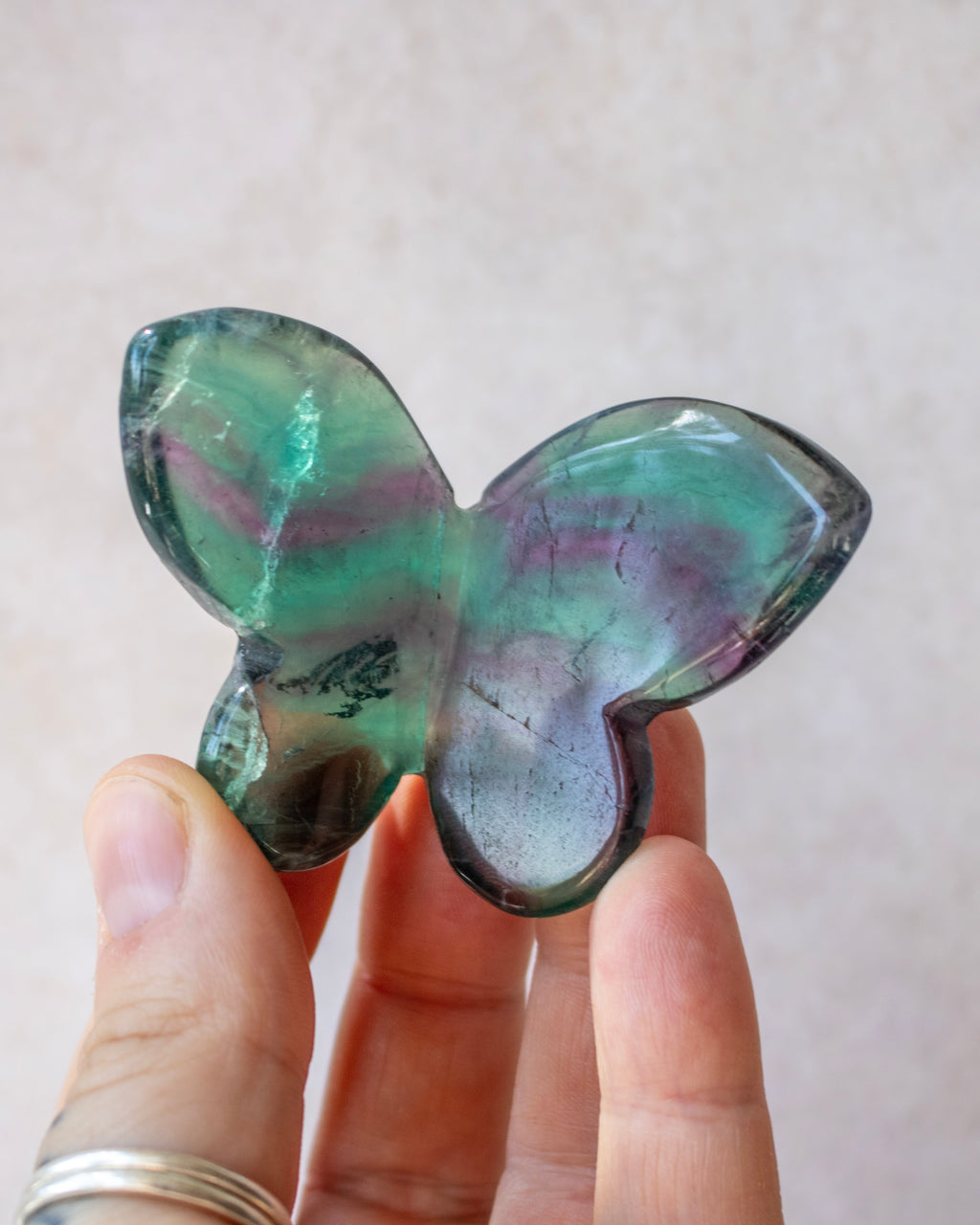 A butterfly-shaped iridescent crystal or gemstone held between fingers. The stone displays shifting colors of green, purple, and blue, with a smooth polished finish.