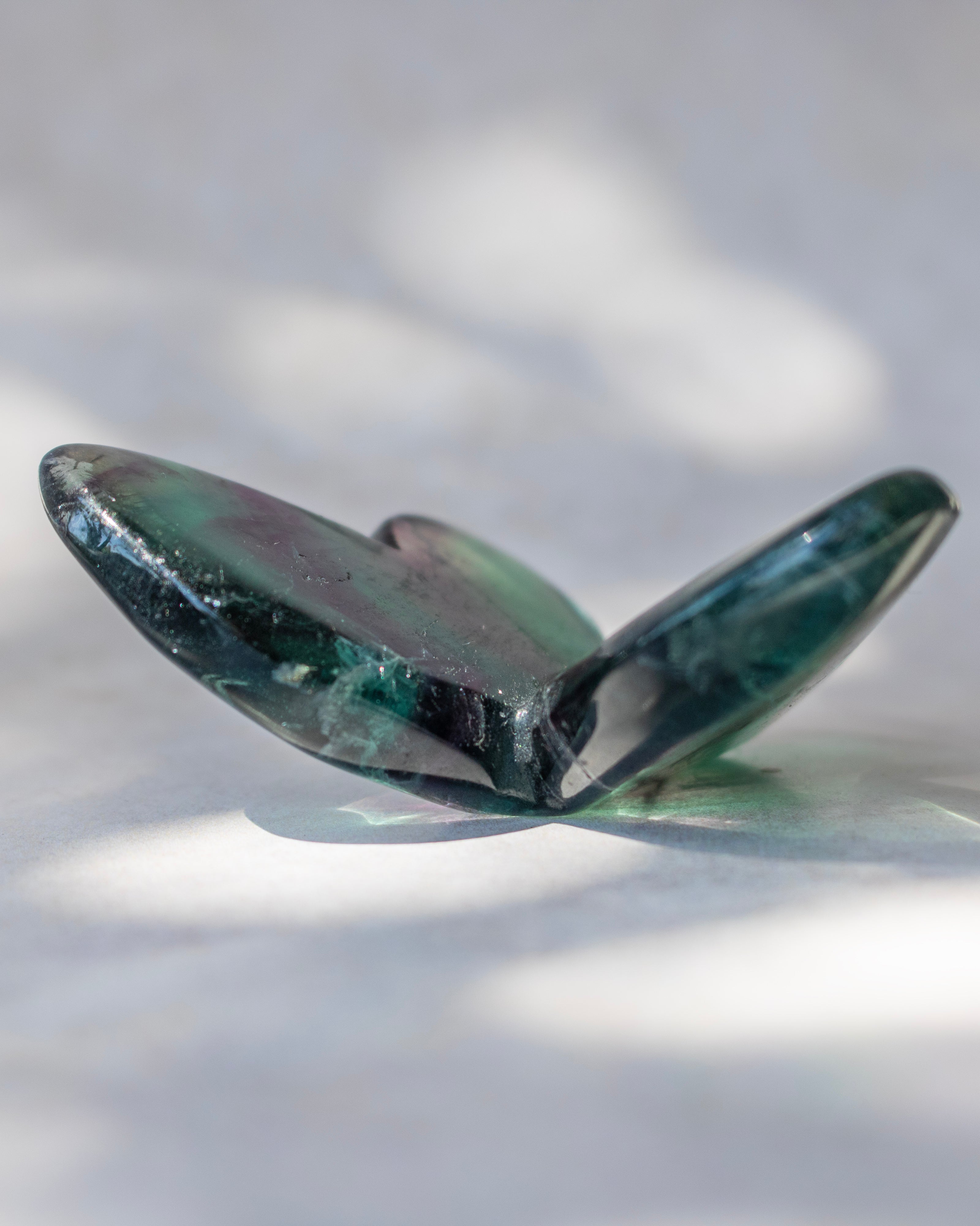 A polished dark green gemstone crystal with a butterfly-like shape, resting on a white surface with soft dappled shadows.
