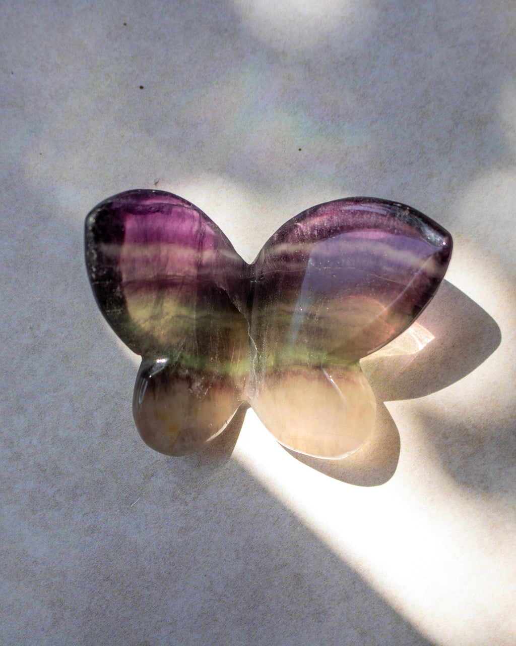 A butterfly-shaped polished gemstone with purple, green, and beige banding, resting on a light gray surface with dappled sunlight and shadows.