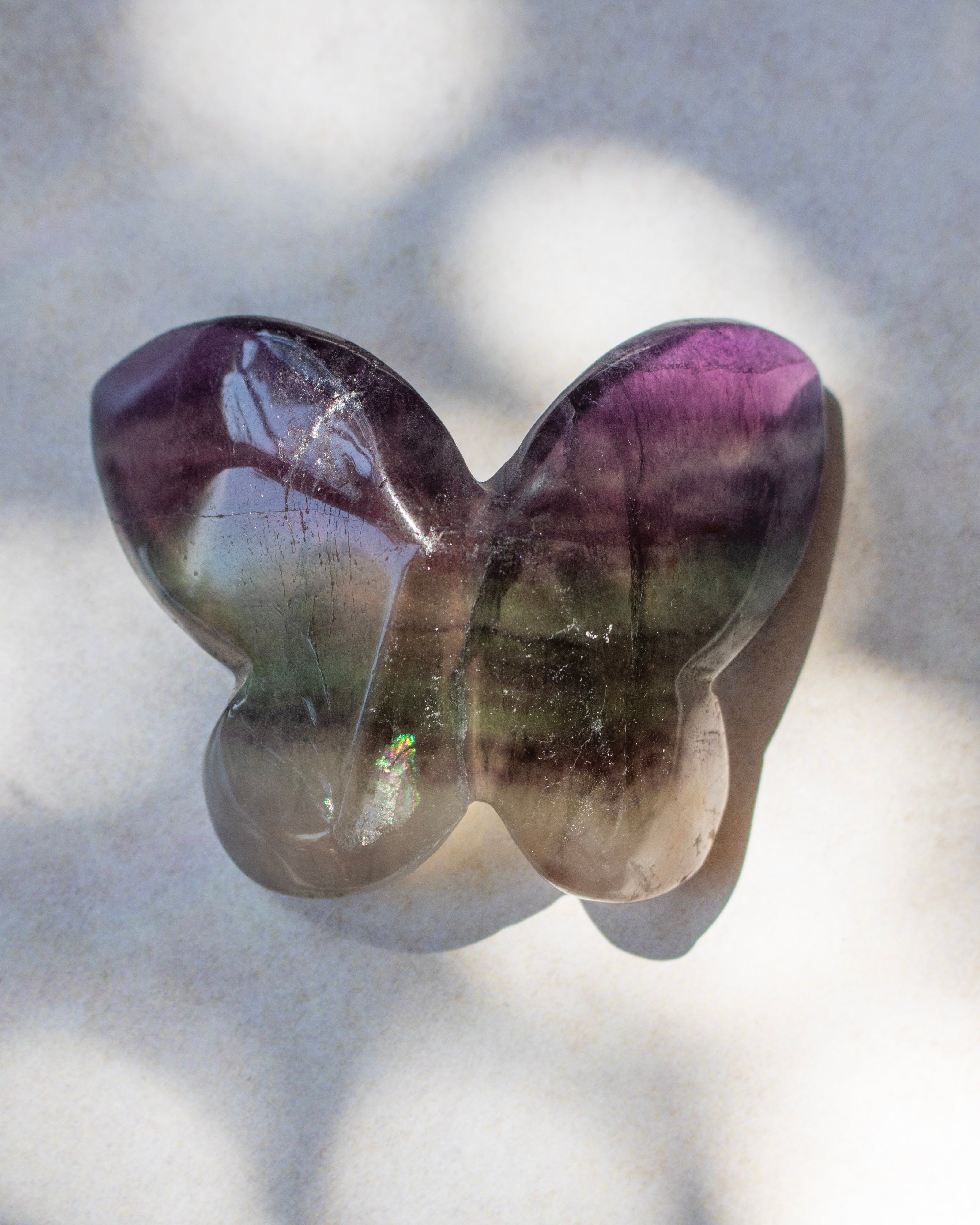 A butterfly-shaped polished gemstone with purple, green, and smoky gray coloration, resting on a light textured surface with soft shadows.