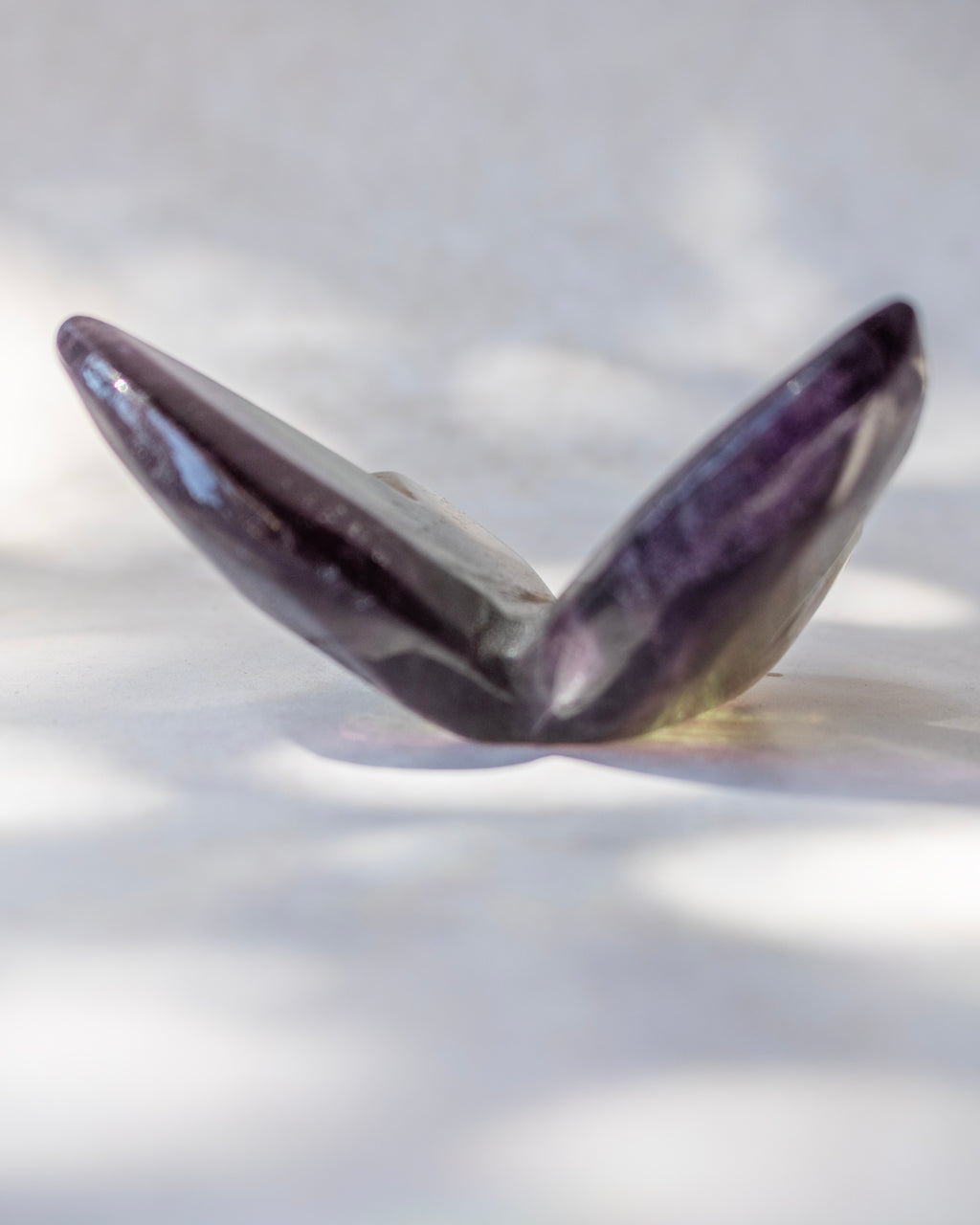 A polished amethyst crystal shell-shaped bead or pendant, showing deep purple and gray tones, resting on a white surface with soft leaf shadows.