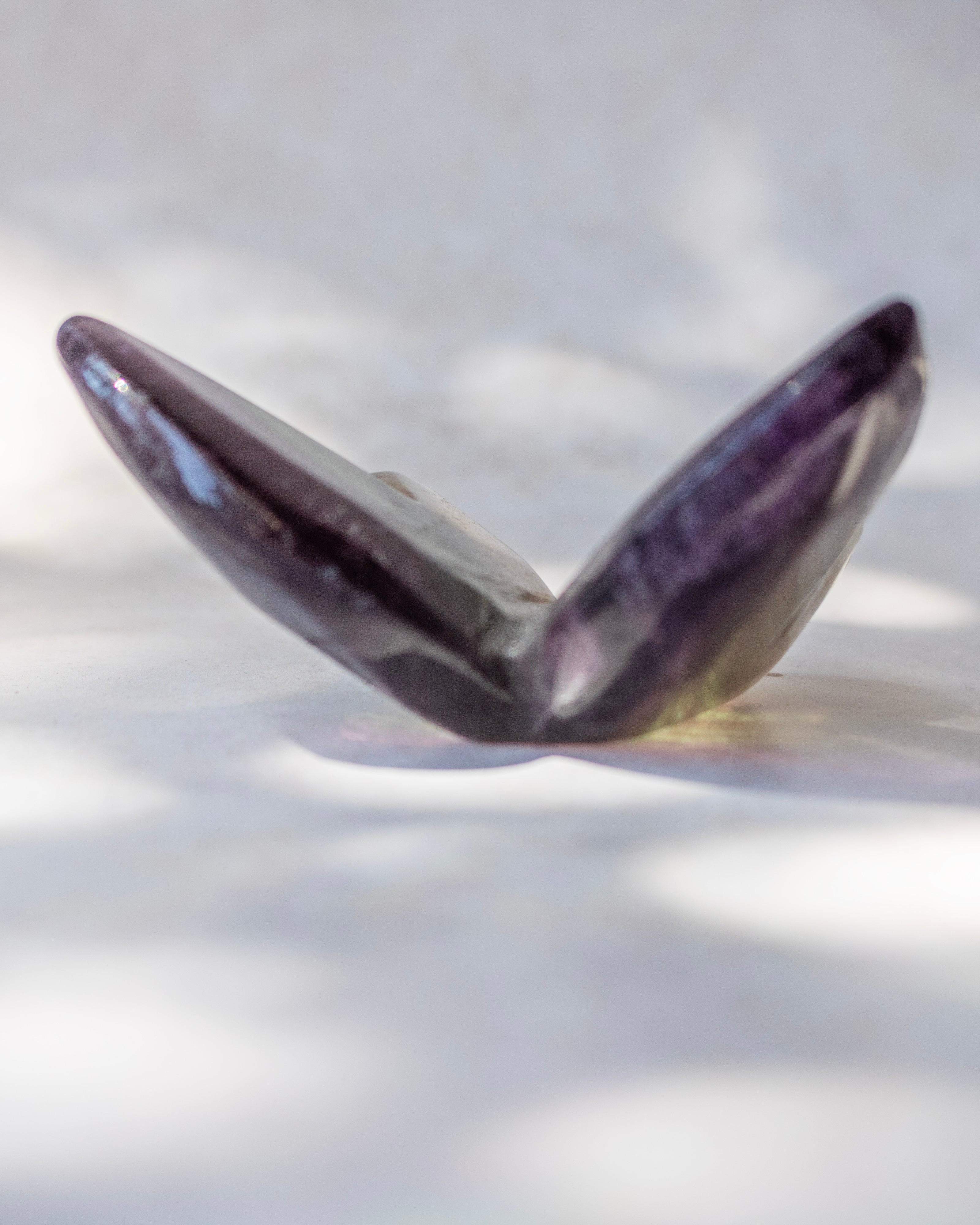 A polished amethyst crystal shell-shaped bead or pendant, showing deep purple and gray tones, resting on a white surface with soft leaf shadows.