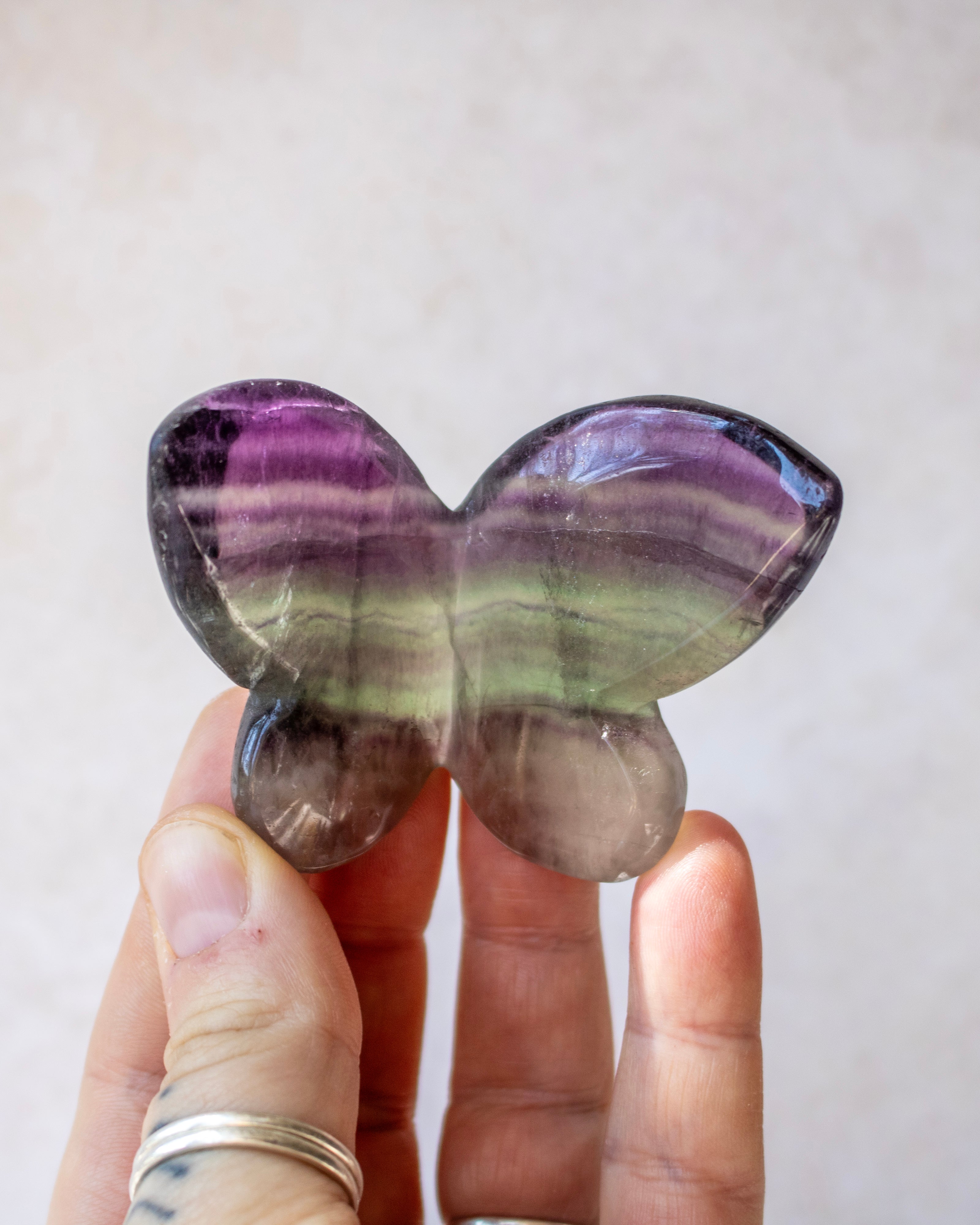 A hand holding a polished butterfly-shaped crystal stone with purple, green, and gray banding, set against a soft beige background.