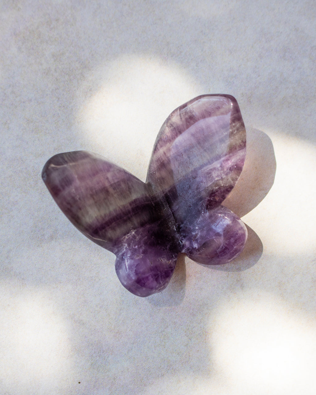 A butterfly-shaped crystal, featuring natural purple and lavender banding, resting on a light textured surface with soft shadows.