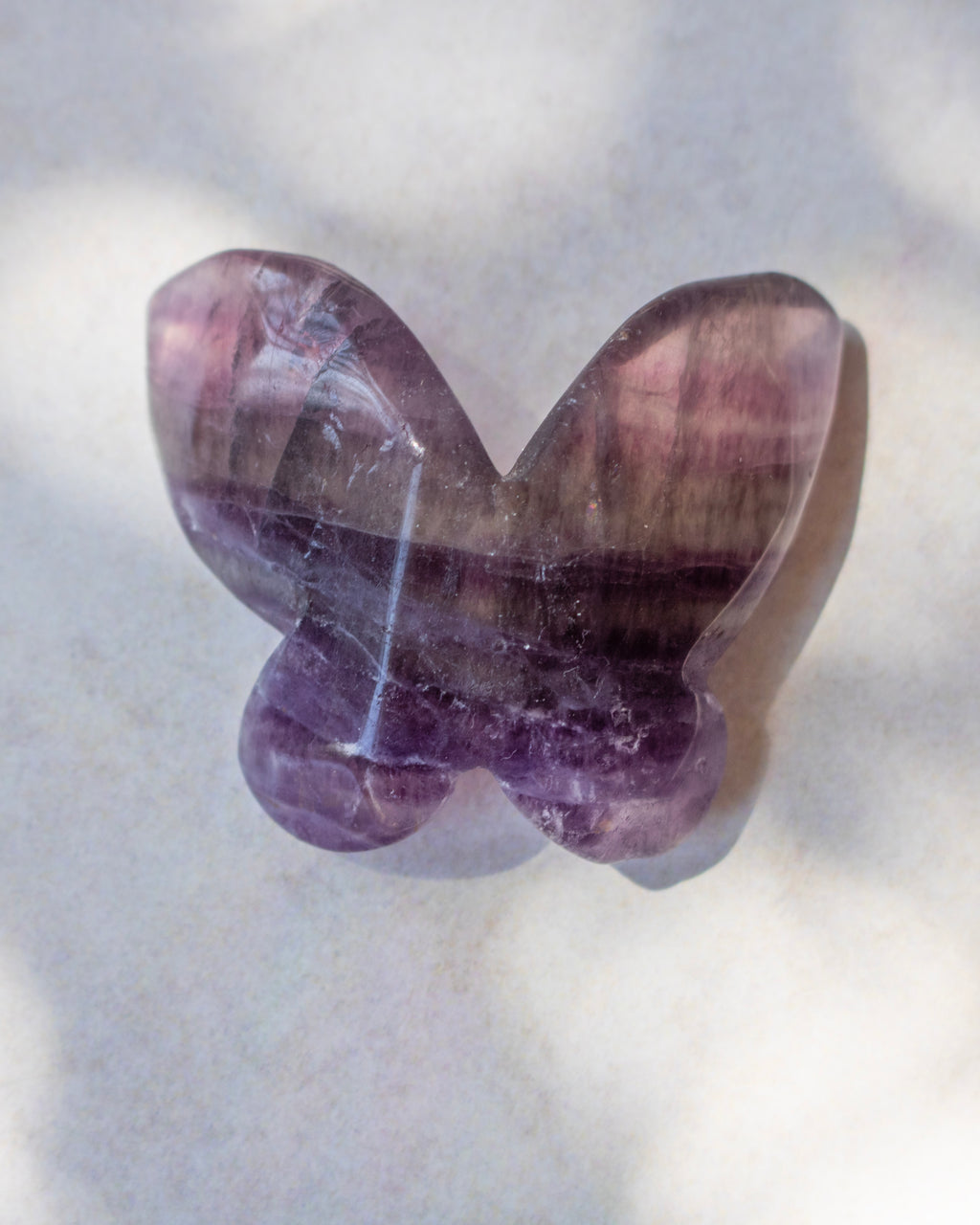 A butterfly-shaped polished gemstone, likely amethyst, resting on a light gray surface with soft shadows.