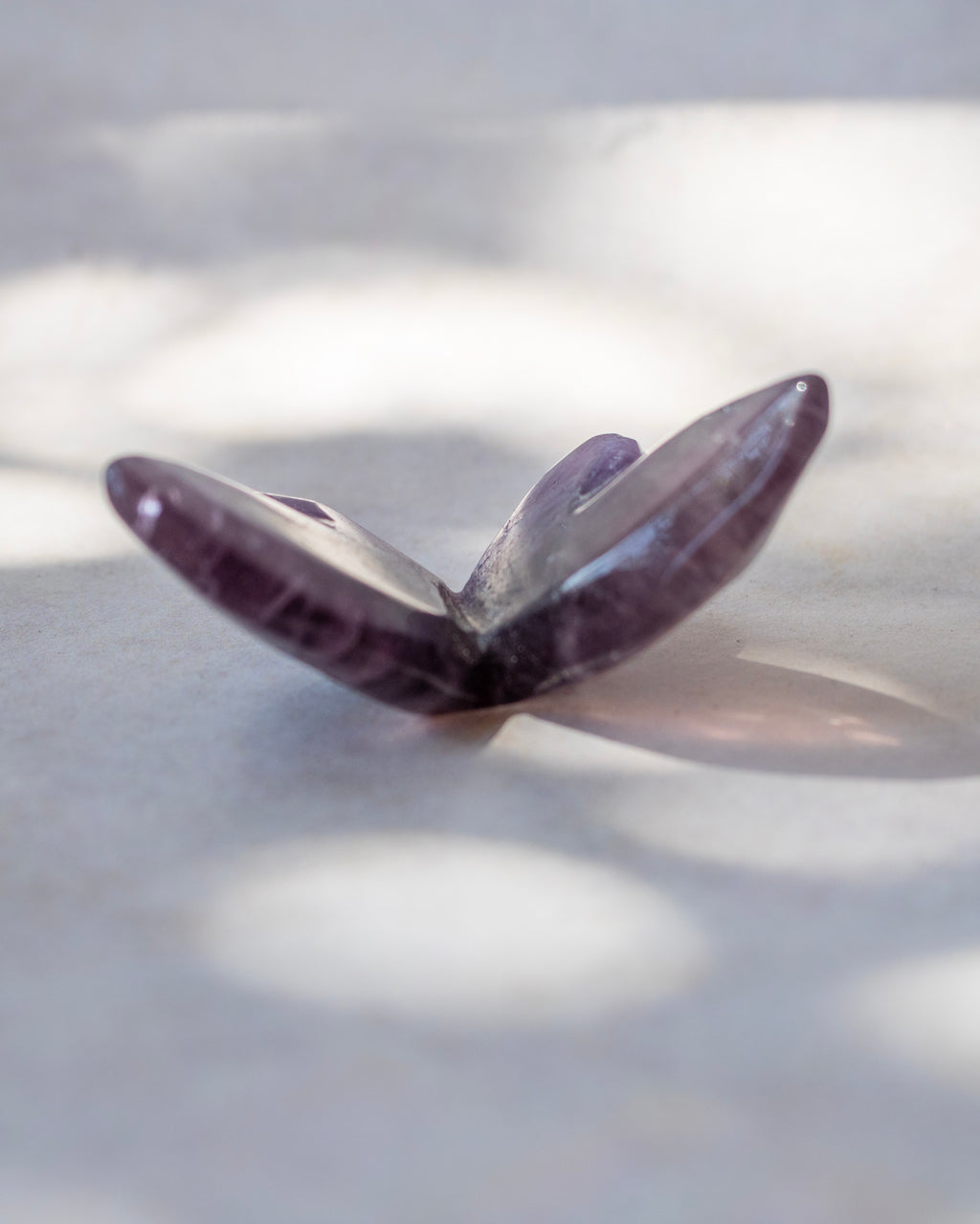 A polished purple crystal butterfly sculpture resting on a light gray surface with soft dappled shadows.