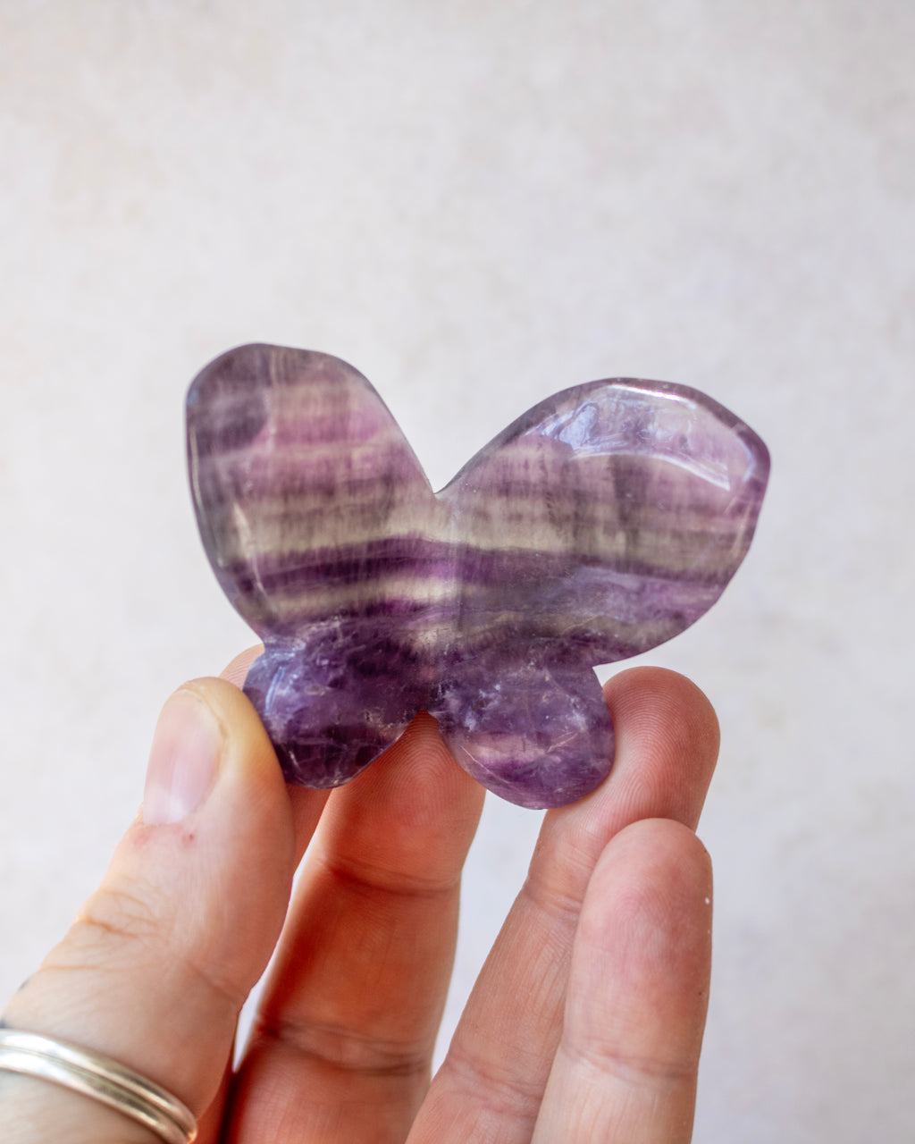 A hand holding a butterfly-shaped polished gemstone with natural purple and white banding, set against a soft, light beige background.