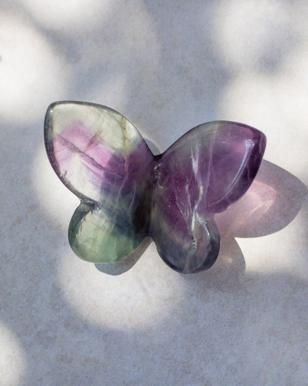 A butterfly-shaped crystal gemstone with a marbled pattern of purple, green, and gray, resting on a light gray textured surface with soft shadows.