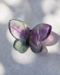 A butterfly-shaped crystal gemstone with a marbled pattern of purple, green, and gray, resting on a light gray textured surface with soft shadows.
