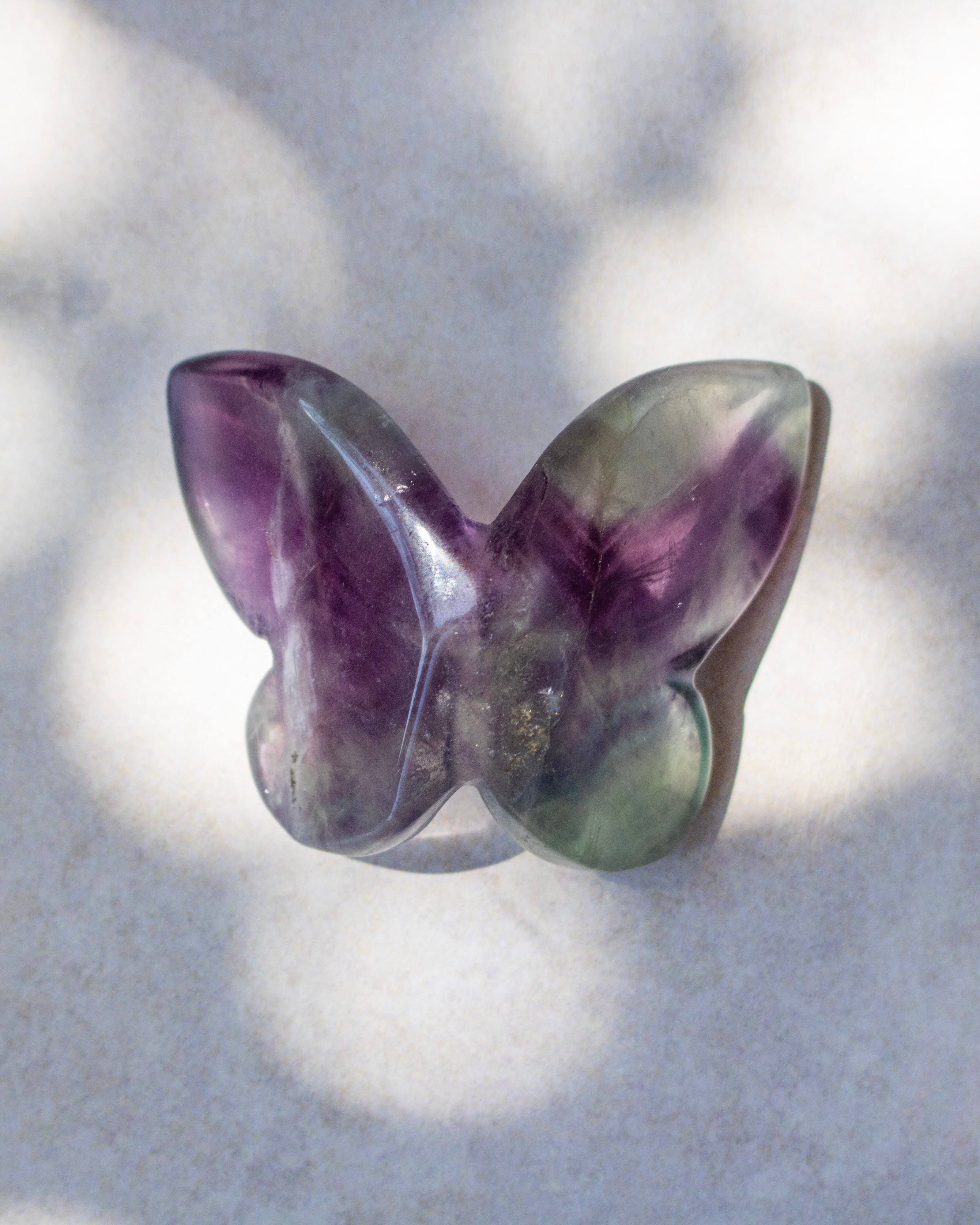 A butterfly-shaped polished gemstone, likely amethyst, featuring purple and green coloration, resting on a light gray textured surface with soft dappled shadows.