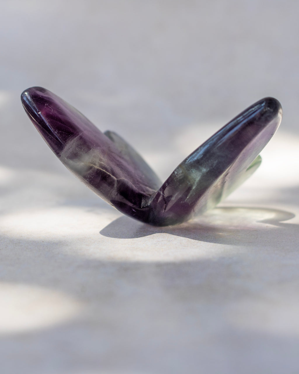 A polished purple amethyst gemstone cut into a butterfly-like shape, displayed on a light gray surface with soft shadows.