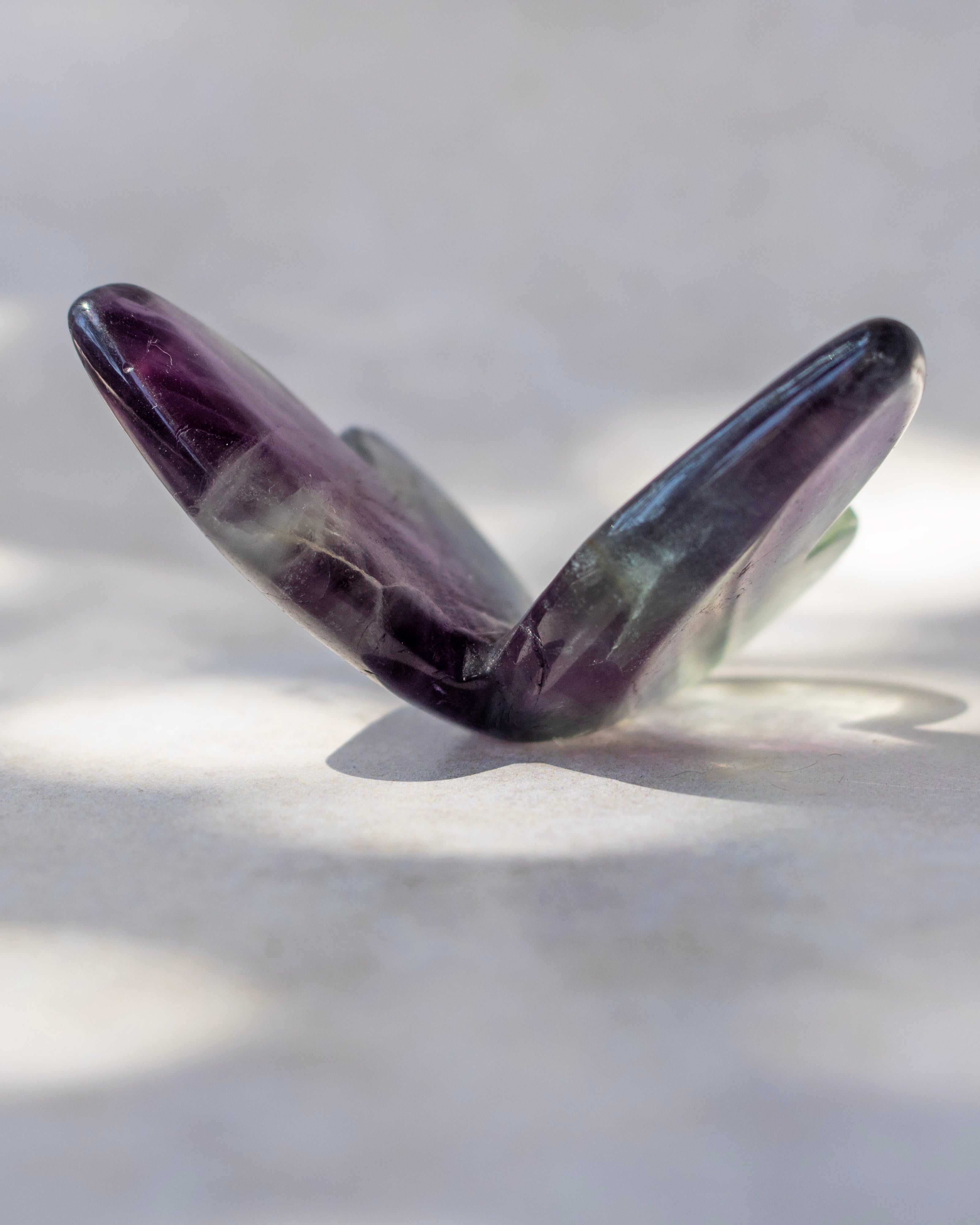 A polished purple amethyst gemstone cut into a butterfly-like shape, displayed on a light gray surface with soft shadows.