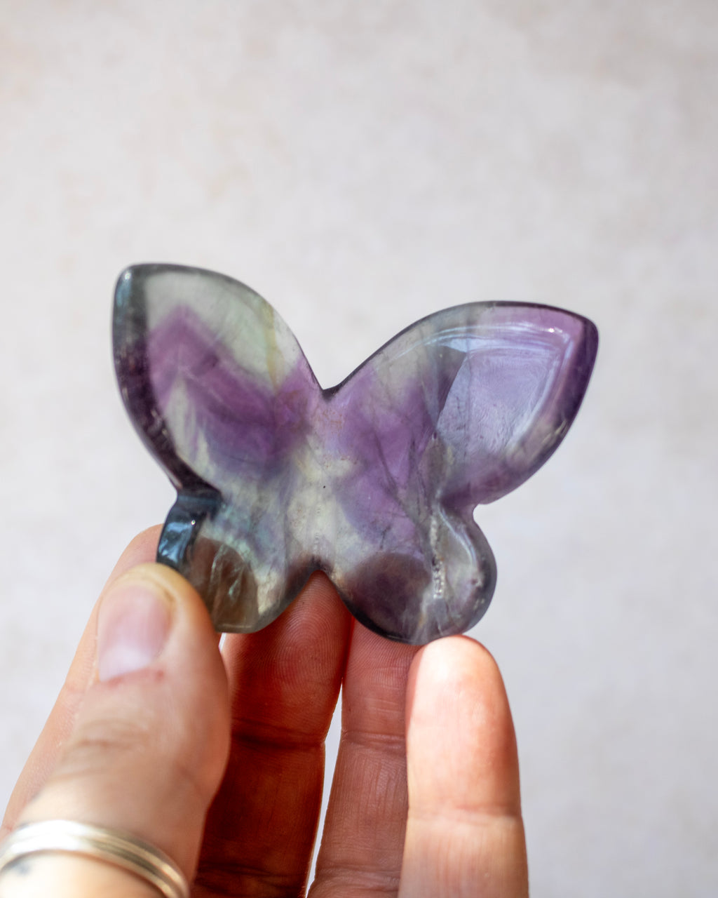 A hand holding a butterfly-shaped polished gemstone with purple, gray, and white marbled patterns against a soft beige background.