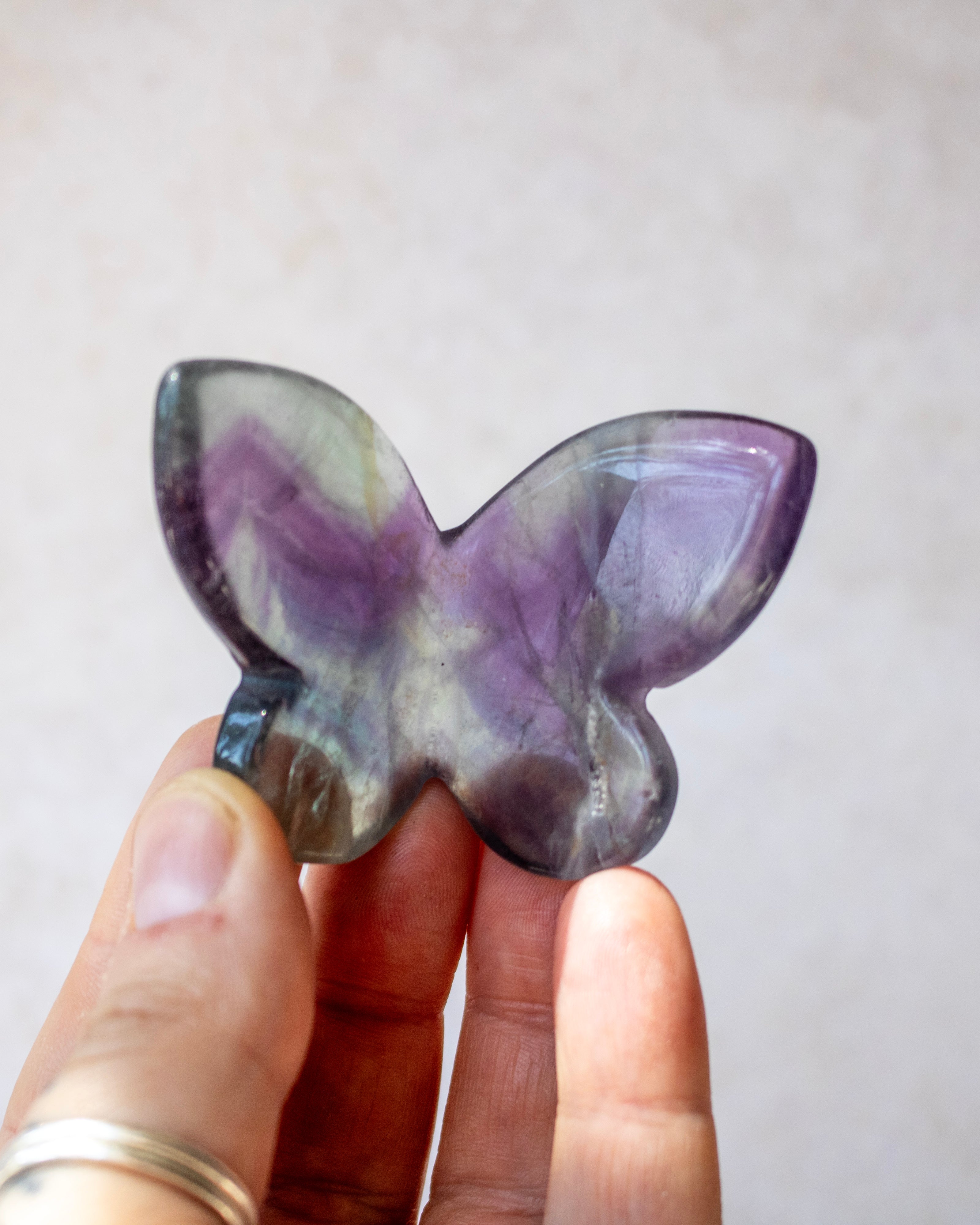 A hand holding a butterfly-shaped polished gemstone with purple, gray, and white marbled patterns against a soft beige background.