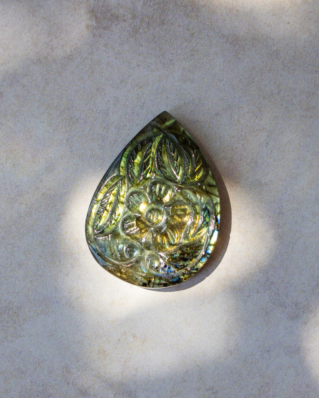 A teardrop-shaped polished gemstone pendant featuring an embossed floral pattern, displaying a shimmering olive green and gold iridescent finish, resting on a textured light gray stone surface.