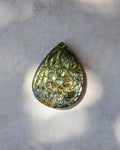 A teardrop-shaped polished gemstone pendant featuring an embossed floral pattern, displaying a shimmering olive green and gold iridescent finish, resting on a textured light gray stone surface.