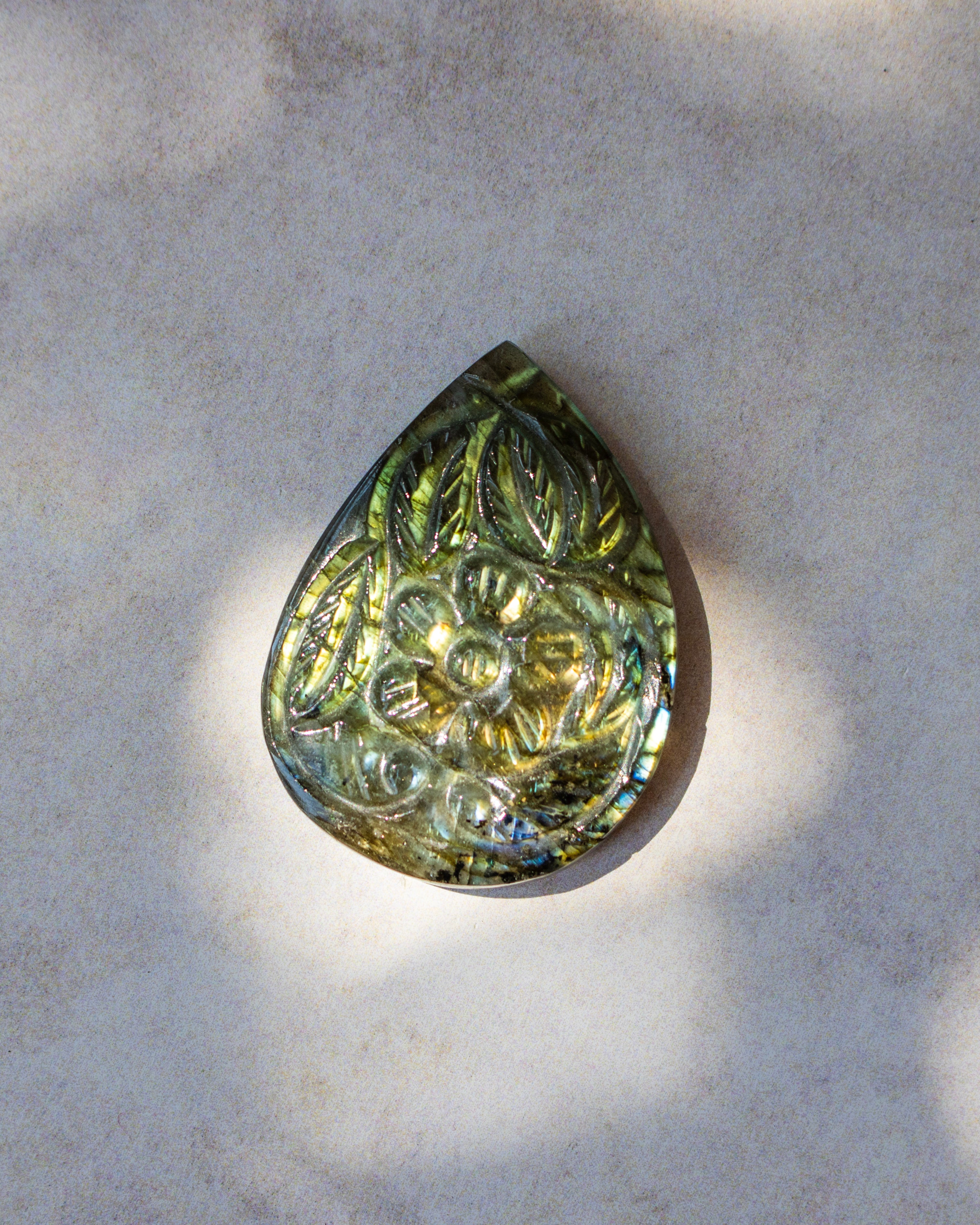 A teardrop-shaped polished gemstone pendant featuring an embossed floral pattern, displaying a shimmering olive green and gold iridescent finish, resting on a textured light gray stone surface.