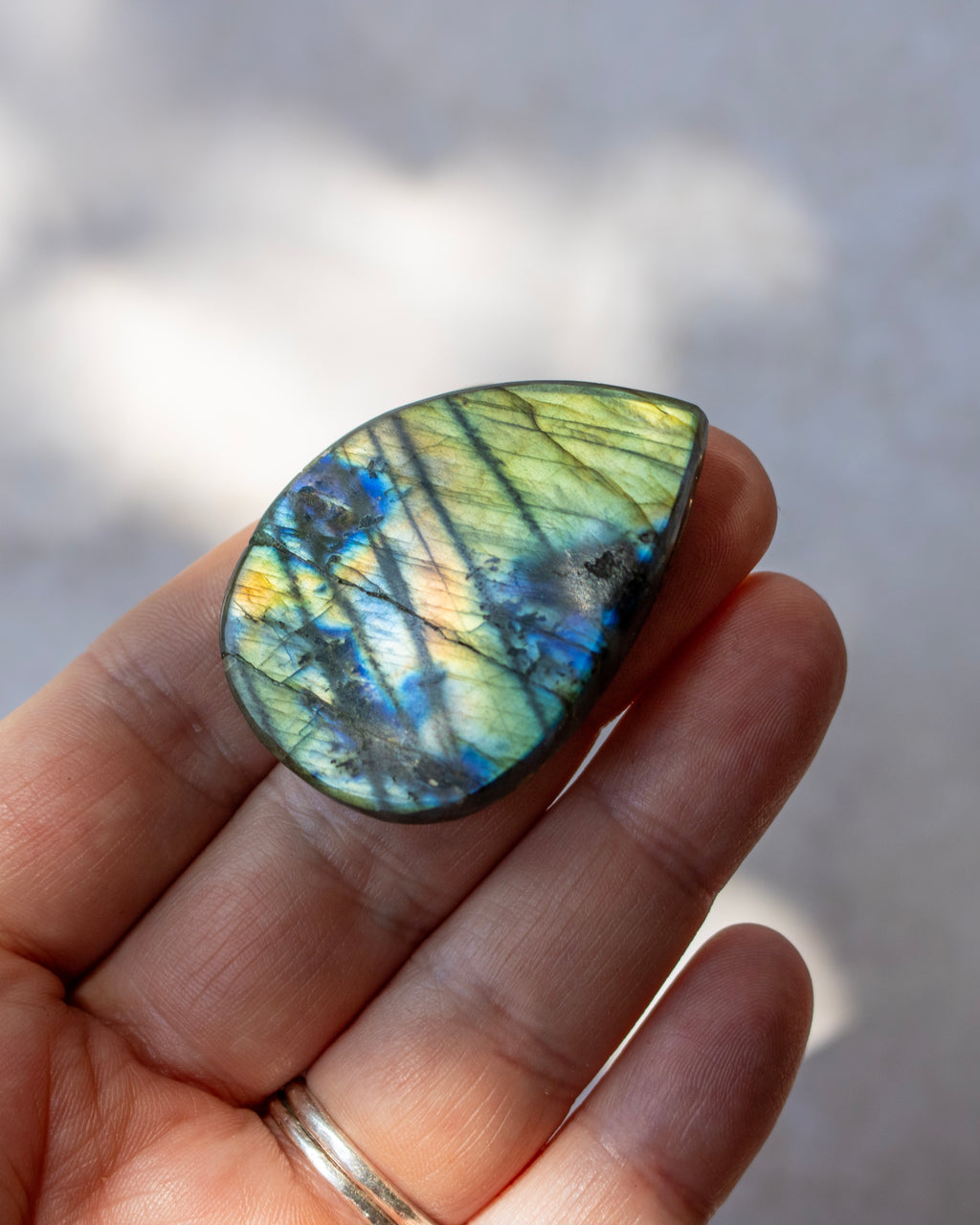 A hand holding a teardrop-shaped labradorite gemstone showing vibrant blue, green, and gold flashes, with a silver ring on the finger against a blurred gray background.