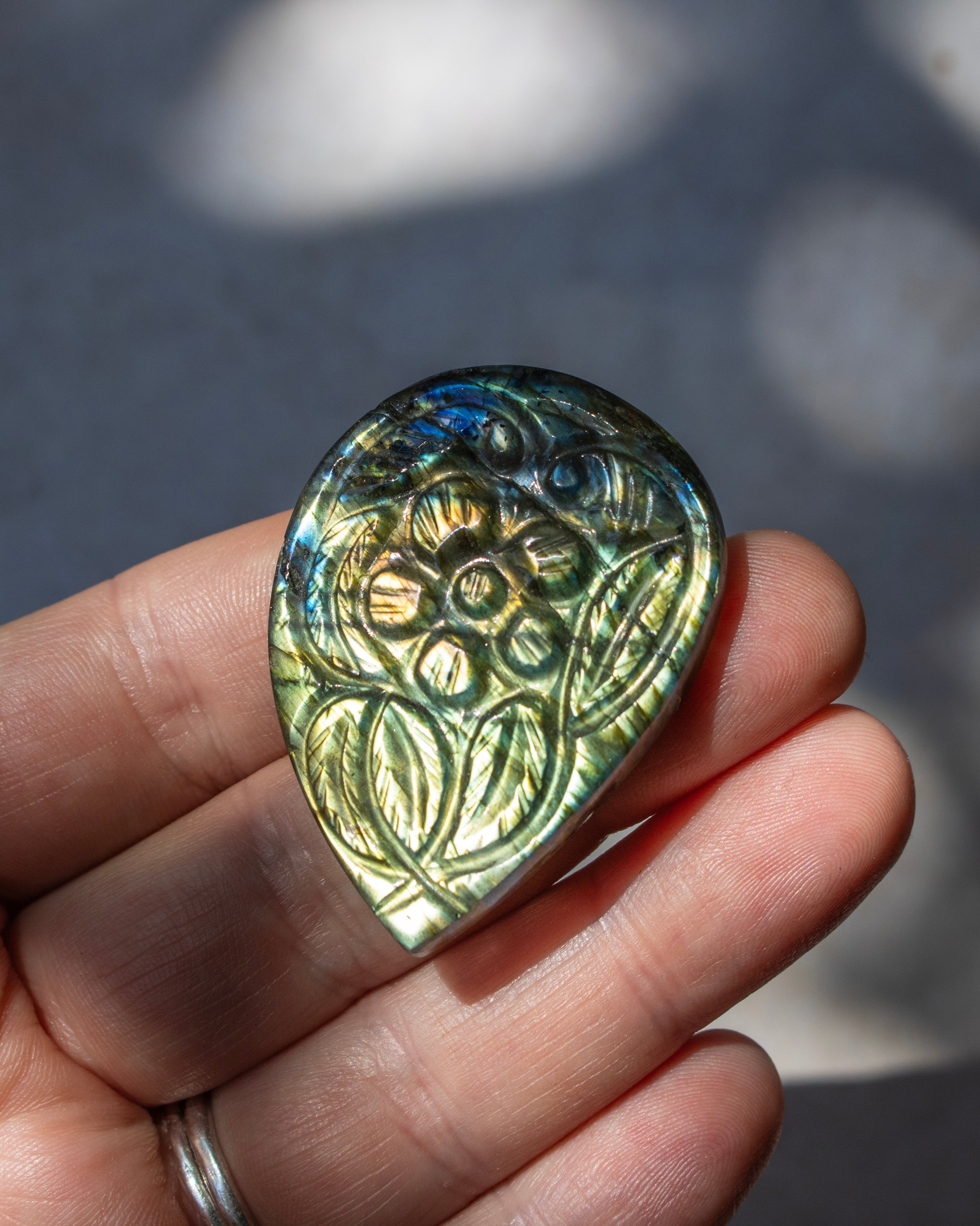 A hand holding a teardrop-shaped polished labradorite gemstone pendant with an engraved floral pattern, showing its characteristic iridescent green and gold color shift.