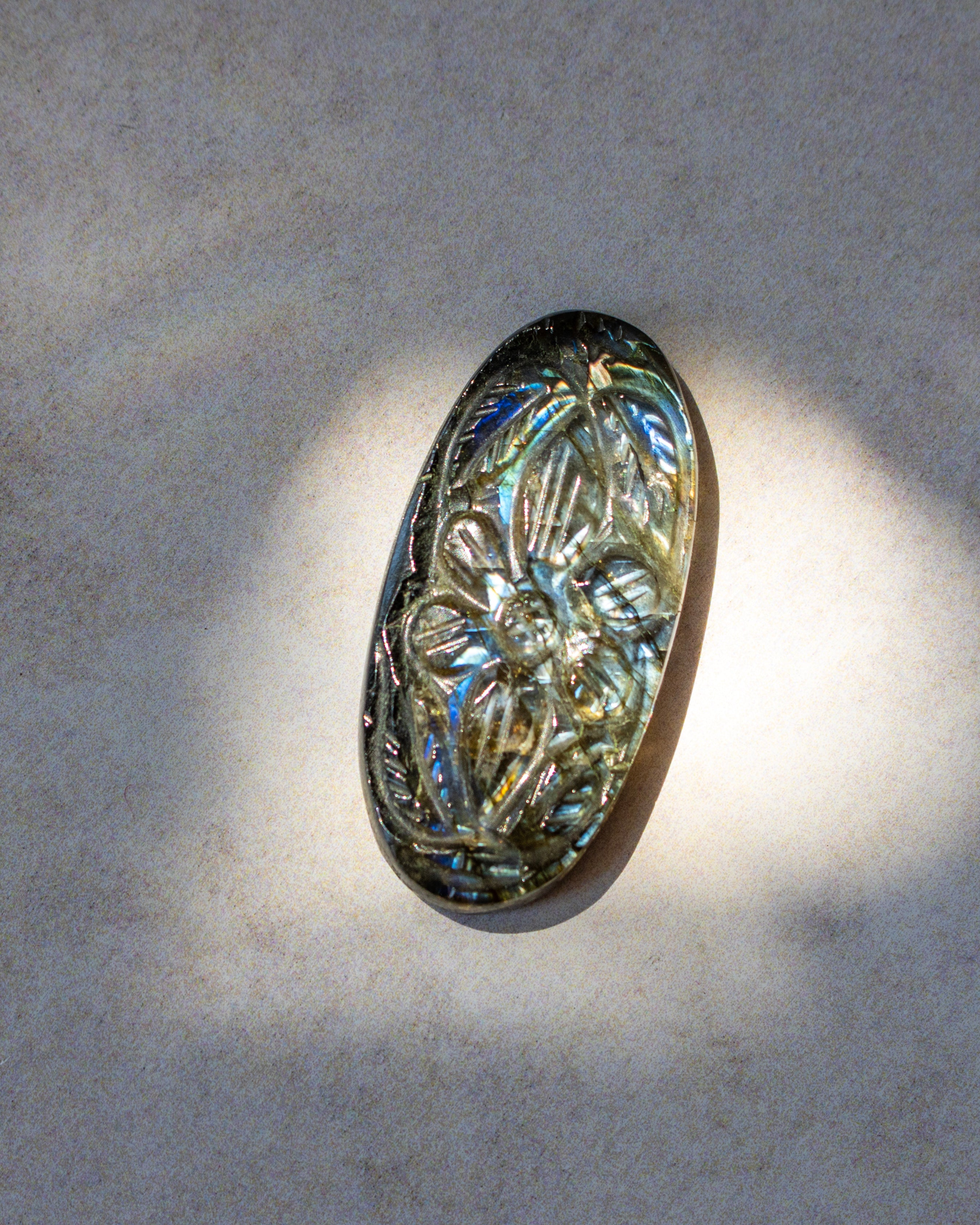An oval-shaped iridescent cabochon with a carved floral pattern, resting on a textured light gray surface under direct sunlight.