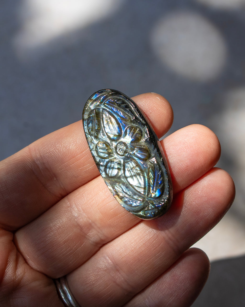 A hand holding an oval polished lapel stone with a floral pattern and iridescent blue-green shimmer, set against a blurred outdoor background.