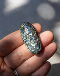 A hand holding an oval polished lapel stone with a floral pattern and iridescent blue-green shimmer, set against a blurred outdoor background.
