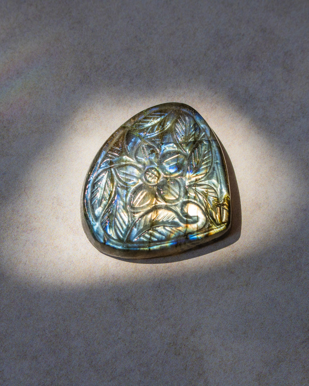 A teardrop-shaped natural labradorite gemstone pendant with an engraved floral pattern, displayed on a textured gray surface under direct spotlighting.