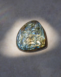 A teardrop-shaped natural labradorite gemstone pendant with an engraved floral pattern, displayed on a textured gray surface under direct spotlighting.