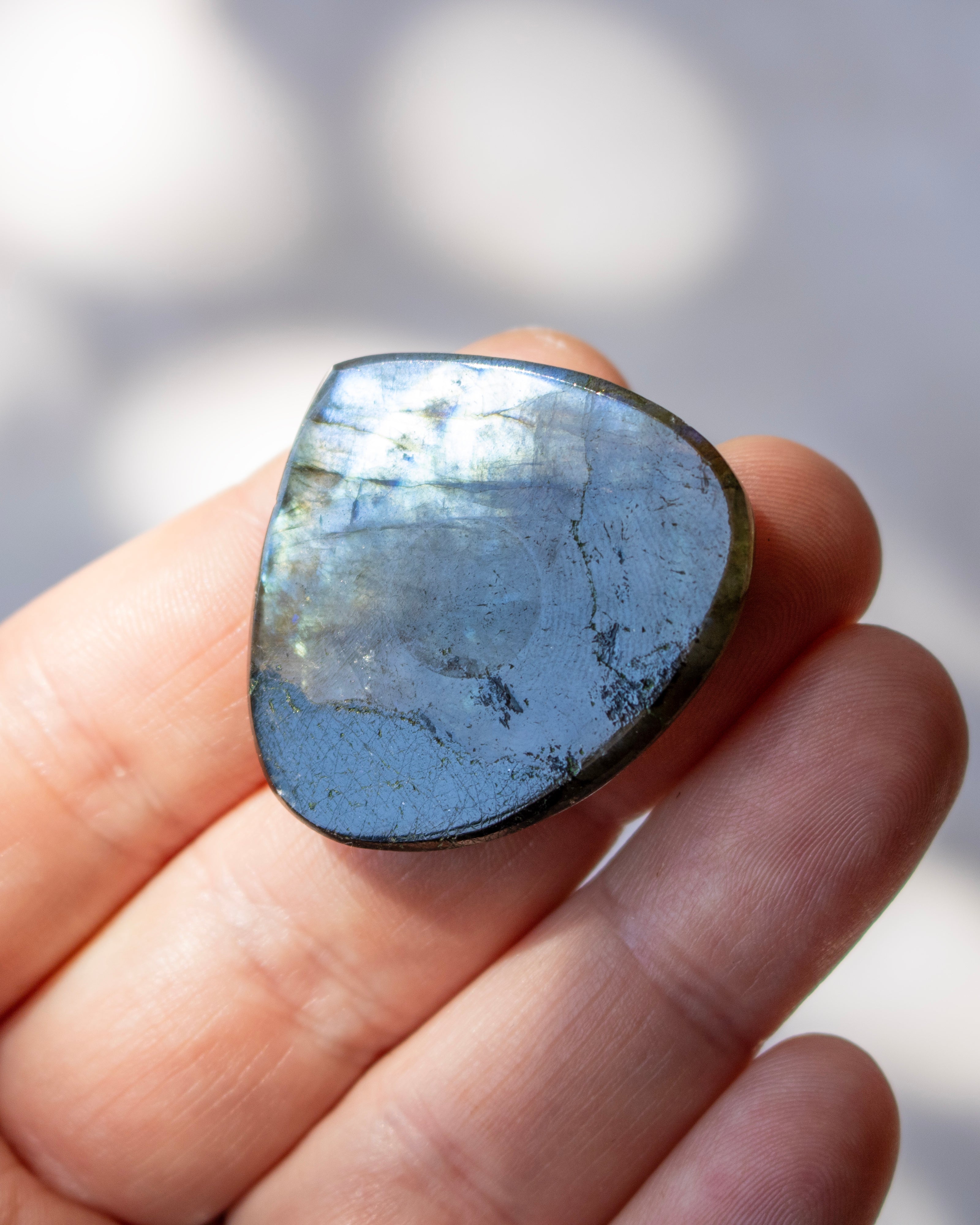 A polished labradorite gemstone cabochon held in a person's hand, showing a shimmering blue-gray color with natural inclusions and a dark border.