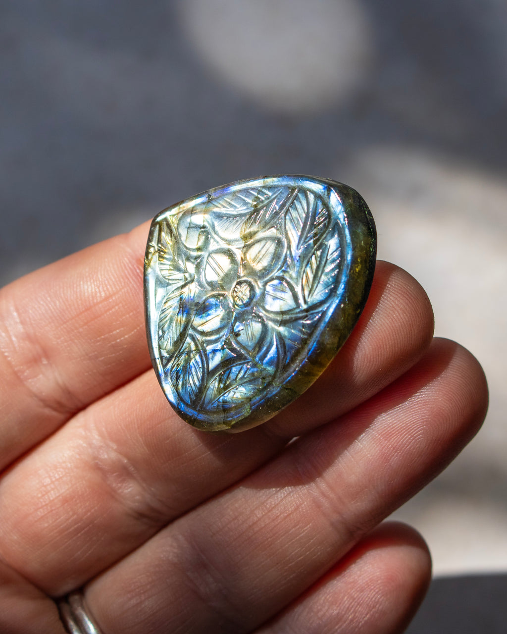 A hand holding a polished iridescent stone cabochon with a floral relief pattern, showing blue, green, and gold shifts under natural light.