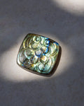 A square lapel pin featuring an embossed floral pattern on a silver-toned base, set with a vibrant iridescent labradorite stone that shifts between green, blue, and gold. The pin is photographed on a neutral gray textured surface with dramatic, direct sunlight casting soft shadows.