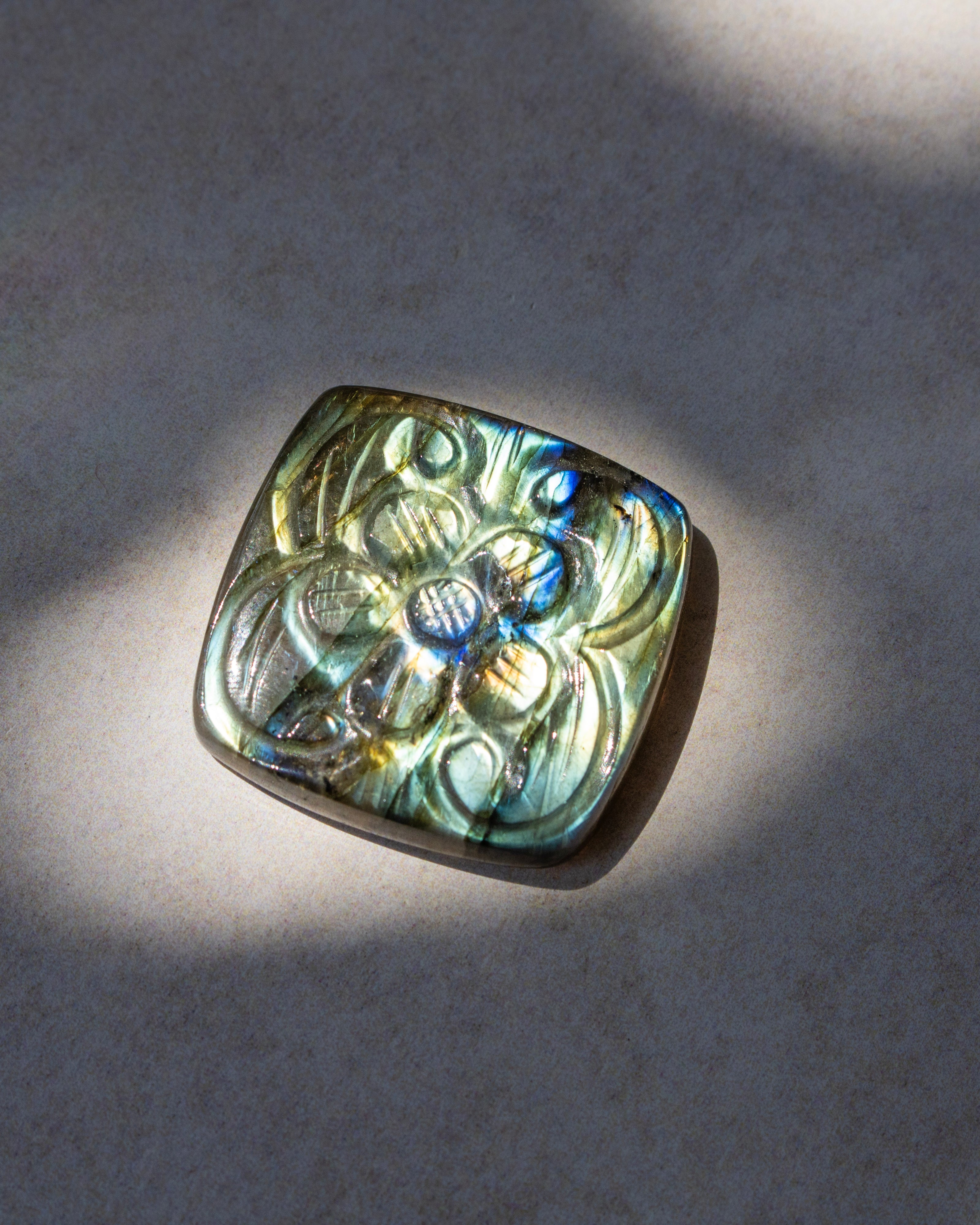A square lapel pin featuring an embossed floral pattern on a silver-toned base, set with a vibrant iridescent labradorite stone that shifts between green, blue, and gold. The pin is photographed on a neutral gray textured surface with dramatic, direct sunlight casting soft shadows.