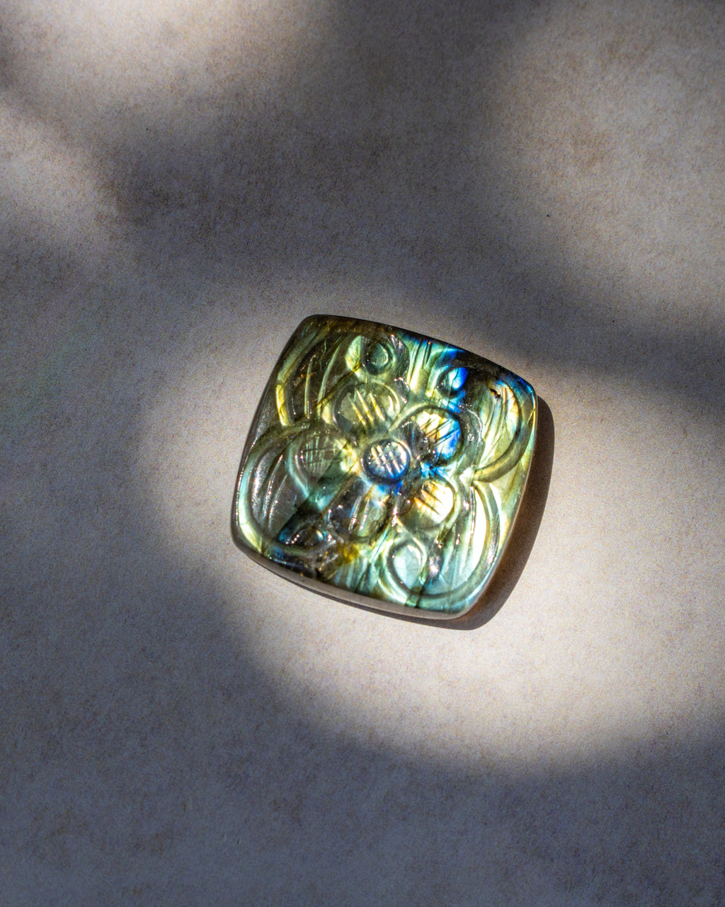 A square pendant with rounded corners featuring an embossed floral pattern, made of iridescent labradorite stone that shifts between green, blue, and gold, resting on a textured beige surface under dappled sunlight.